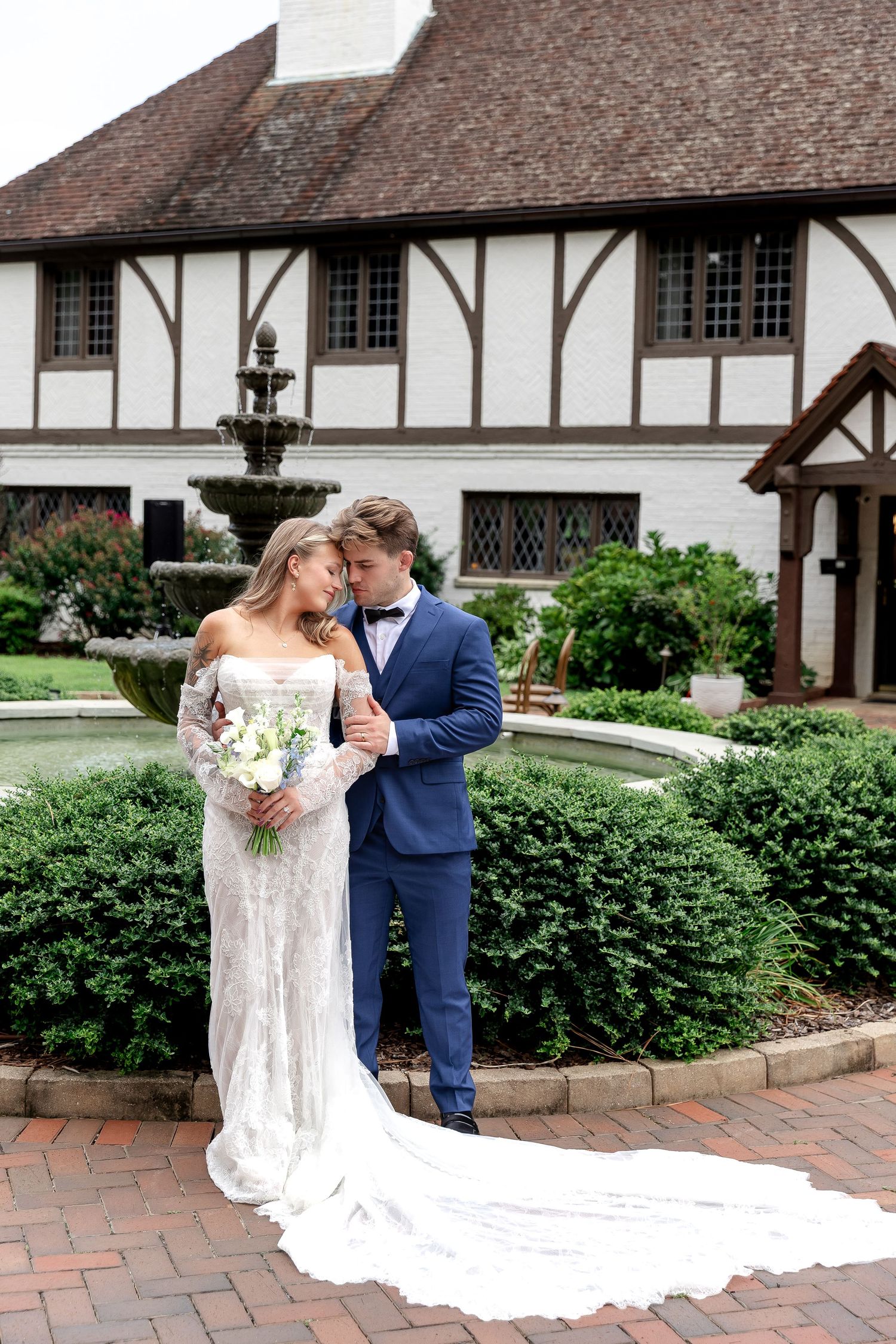 Bride and groom embrace near a fountain outside a Tudor-style mansion on their wedding day.