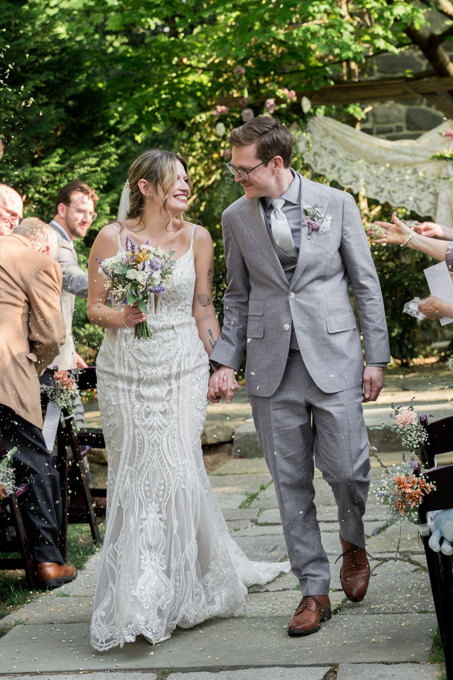 Bride and groom walk down aisle after outdoor wedding ceremony, confetti falling, bride holding wildflower bouquet.
