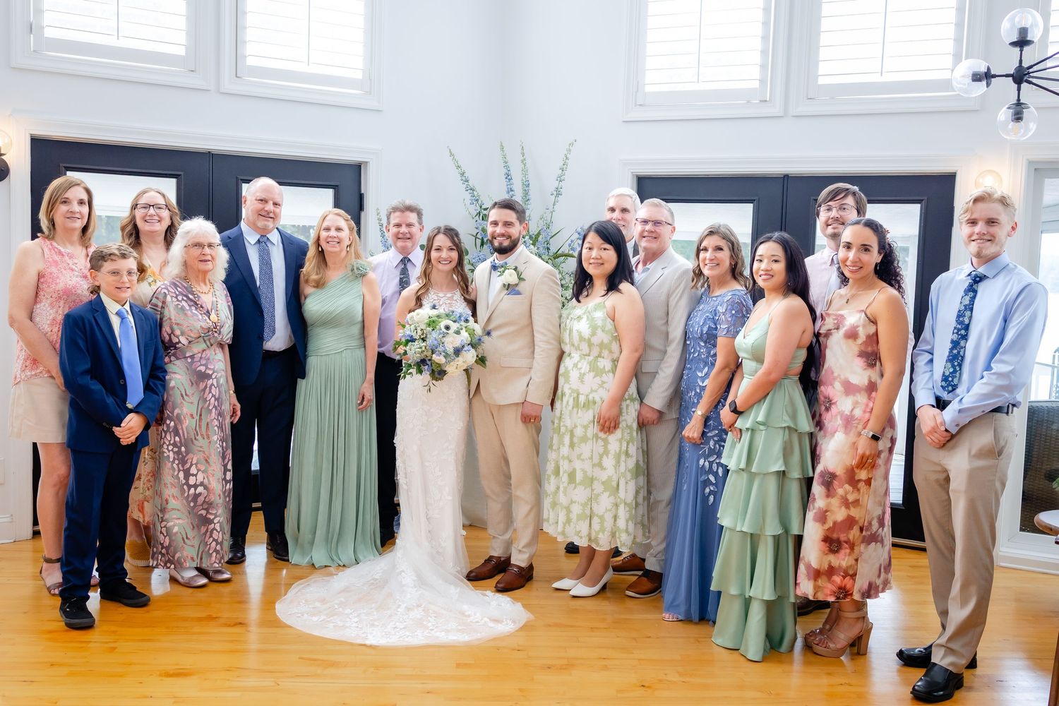 Bride in a white lace gown and groom in a tan suit pose with smiling family and guests in a bright wedding venue with wood floors.