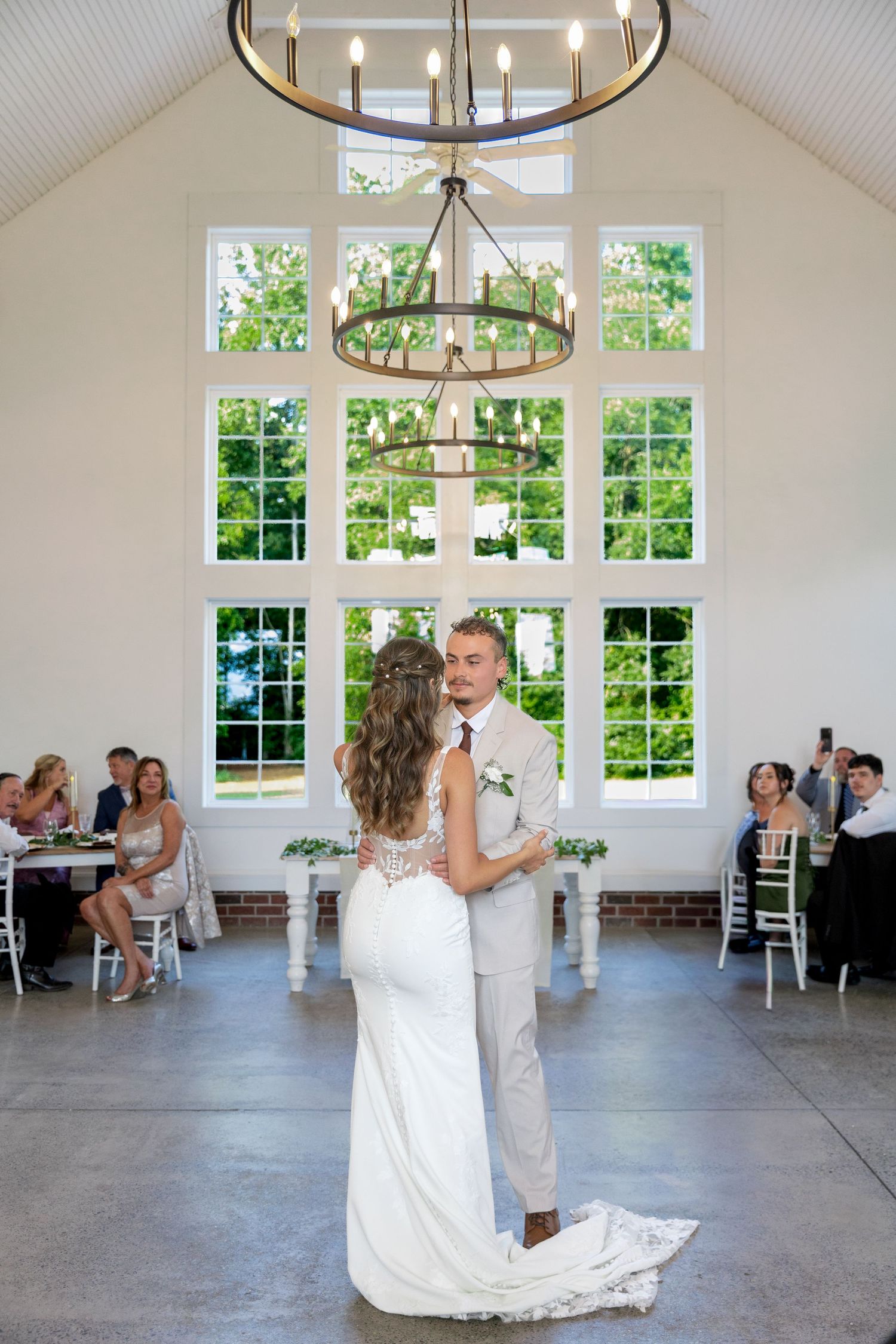Bride and groom share a first dance in a bright reception hall with tall windows, chandeliers, and seated wedding guests looking on.