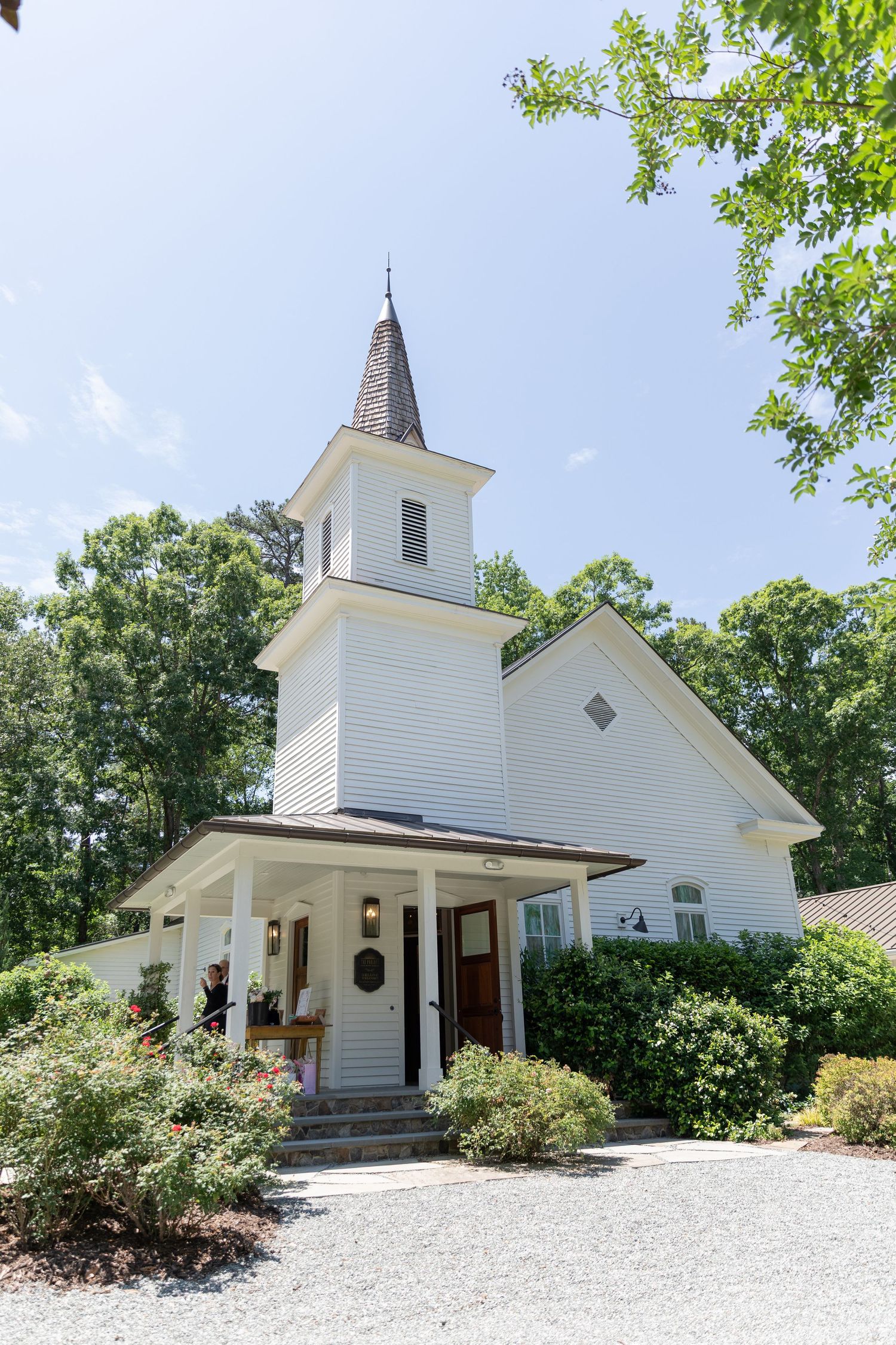 Charming white chapel wedding venue with steeple surrounded by lush trees on sunny day.