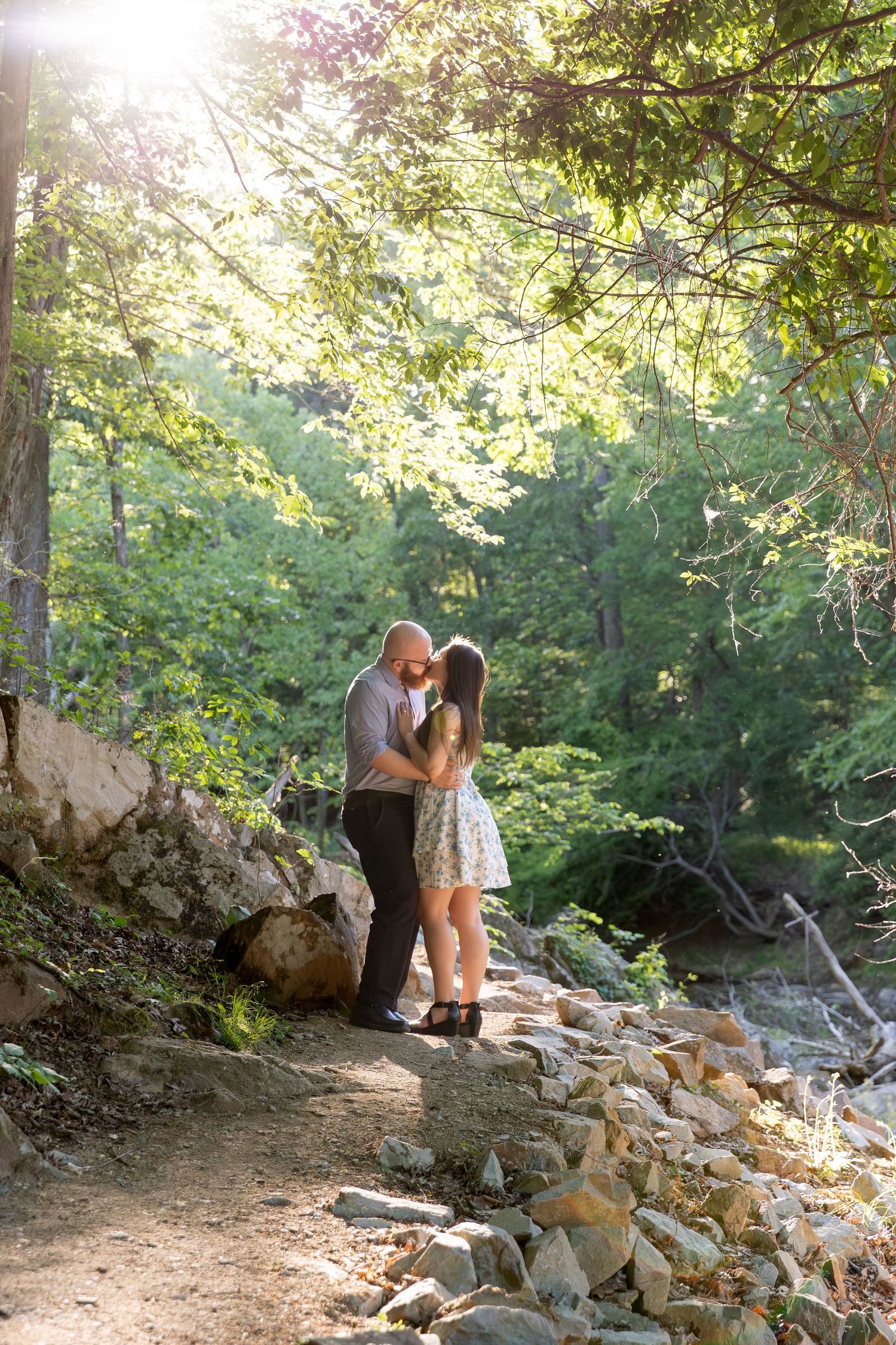 Couple shares a romantic kiss on a rocky forest path surrounded by lush green trees.