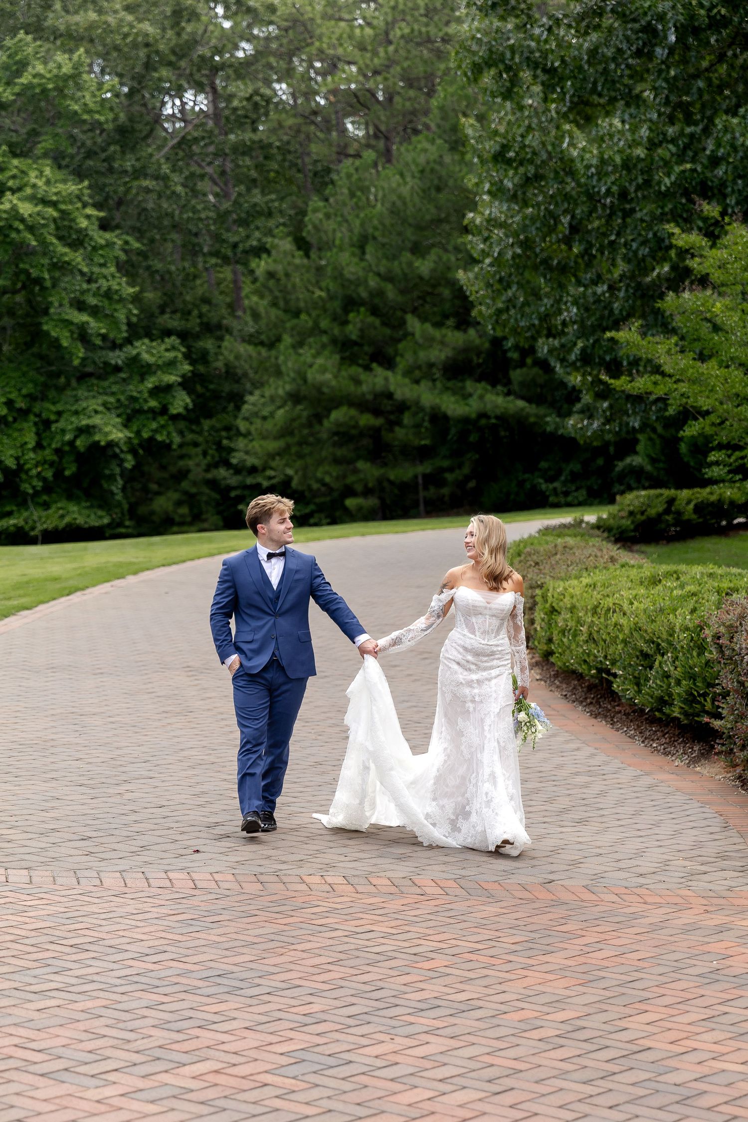Bride and groom walk hand-in-hand on brick path surrounded by lush greenery, bride in lace gown, groom in navy suit.