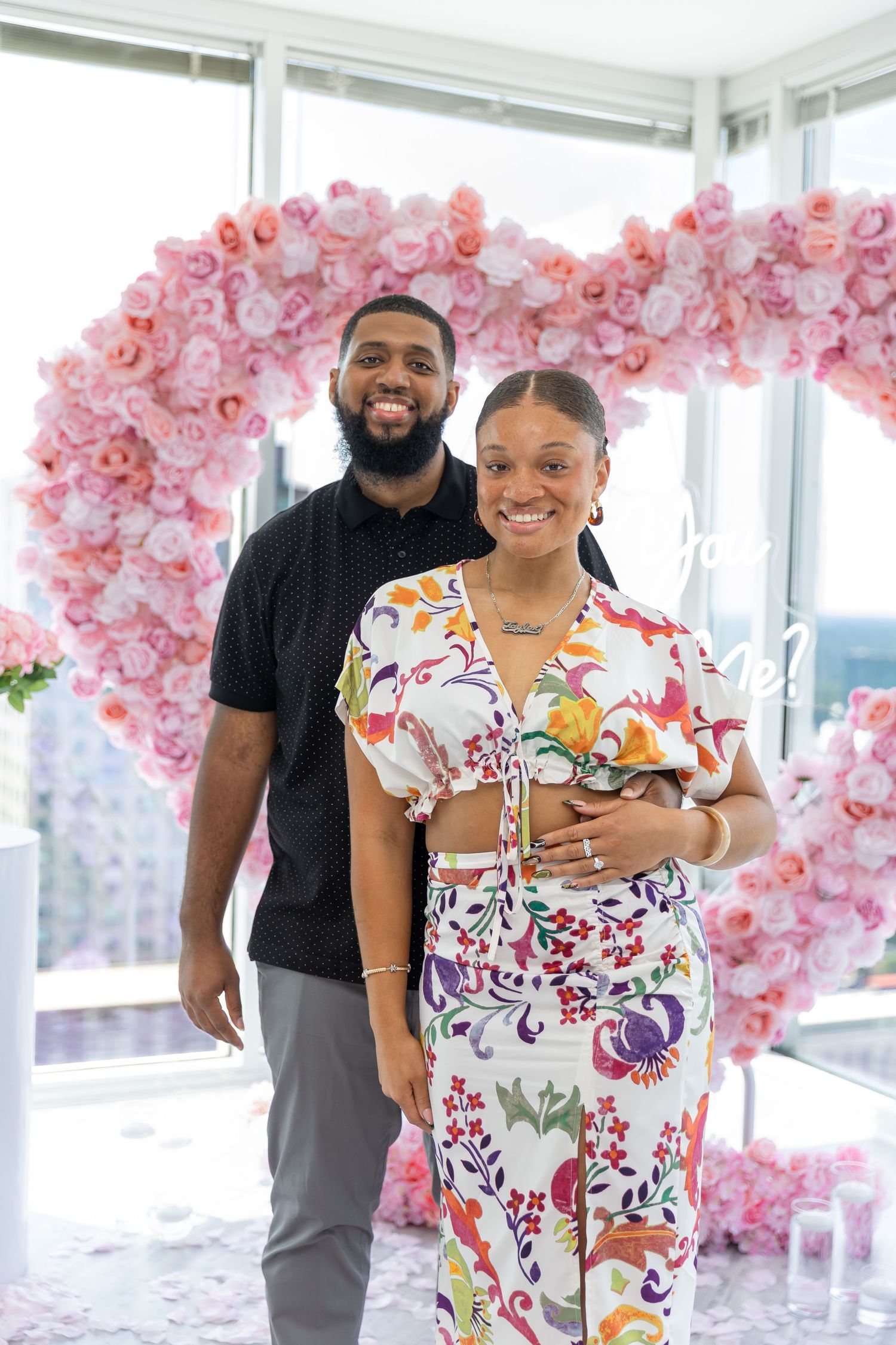 Smiling couple poses in front of a pink floral heart arch, woman in floral outfit, man in black shirt.