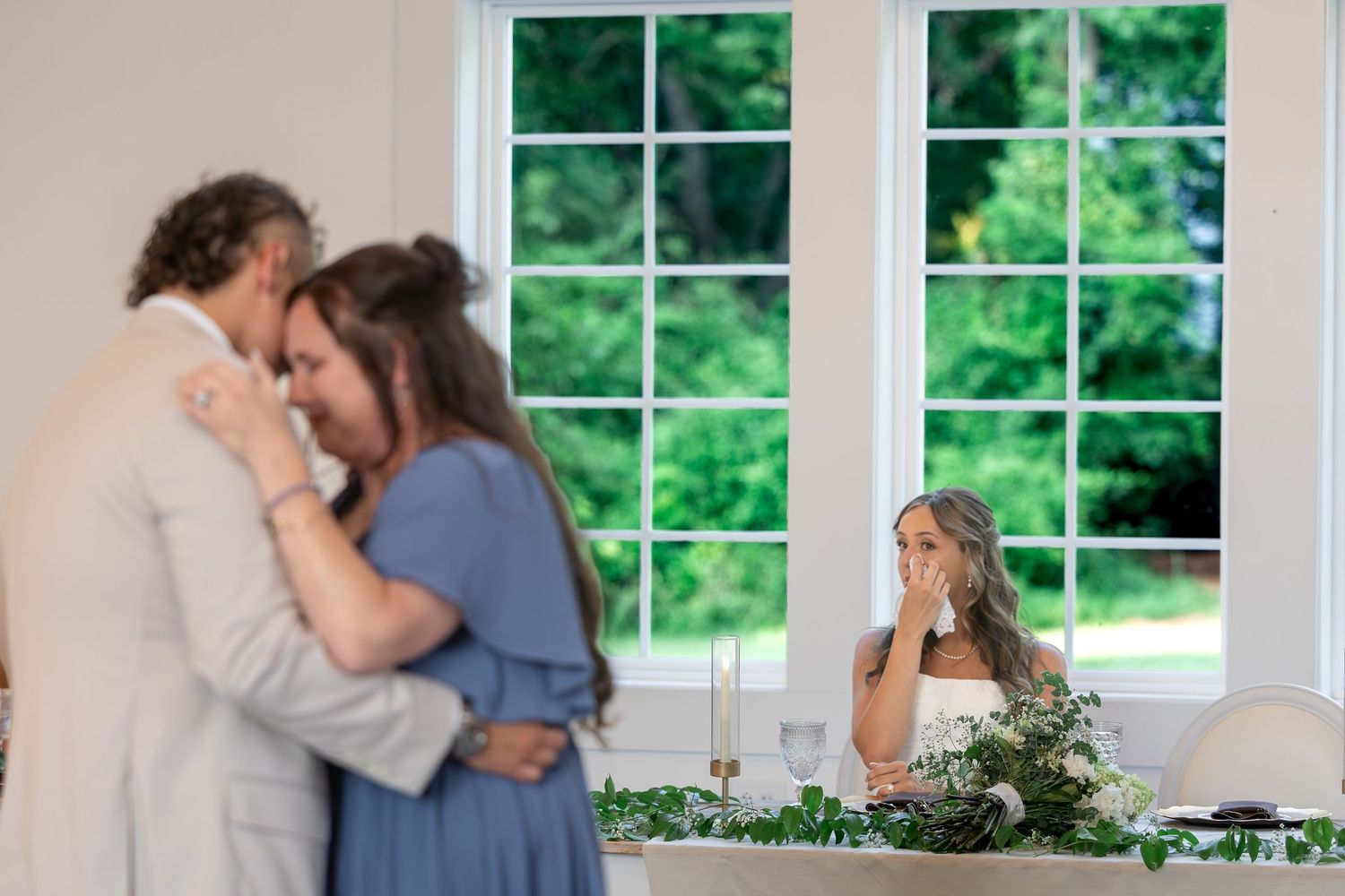 A man in a light suit embraces a woman in a blue dress at a wedding reception while a bride in white wipes away tears nearby.