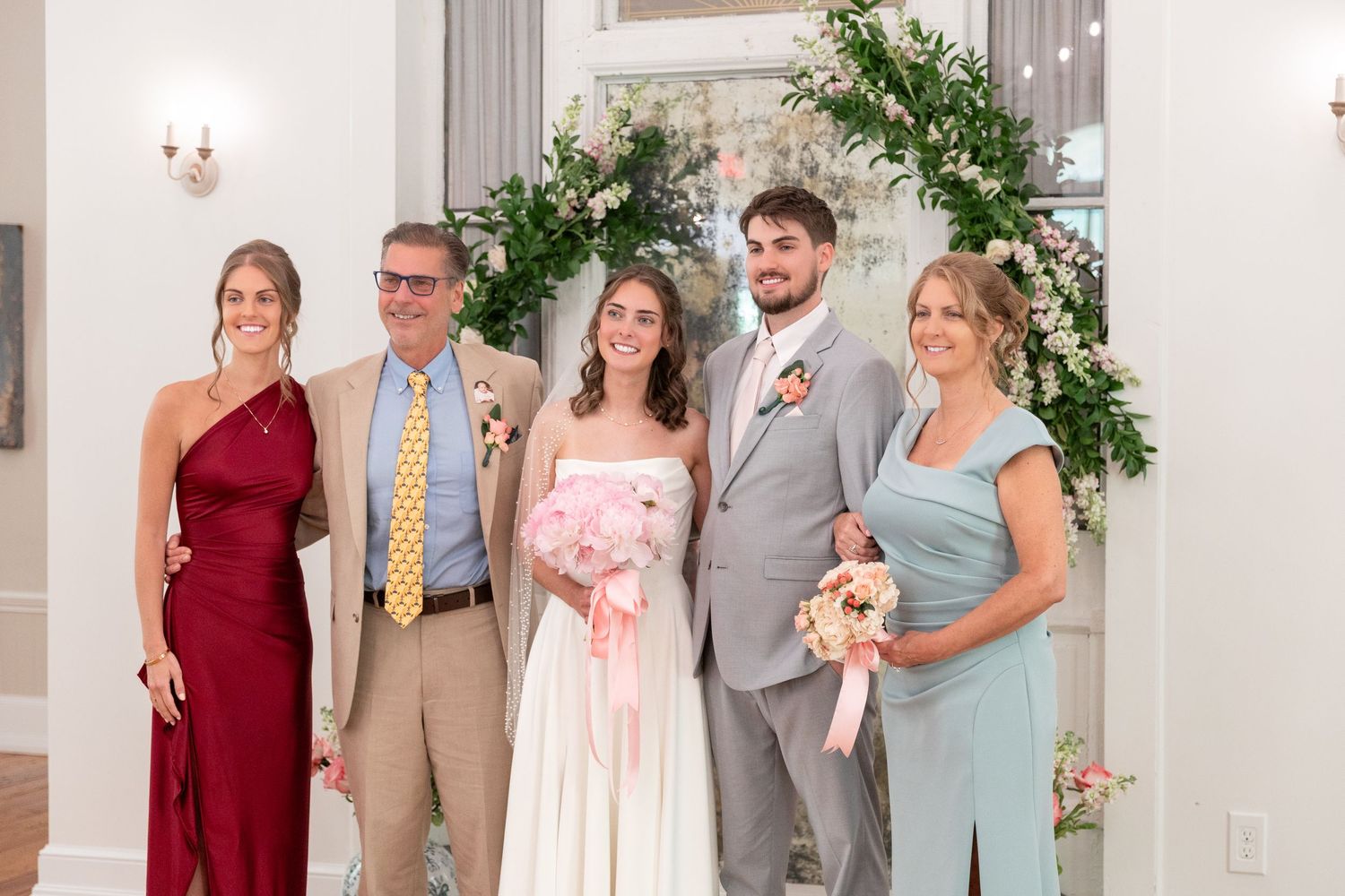 Bride in white gown holding a pink bouquet poses with groom and three family members in formal wedding attire near floral greenery.