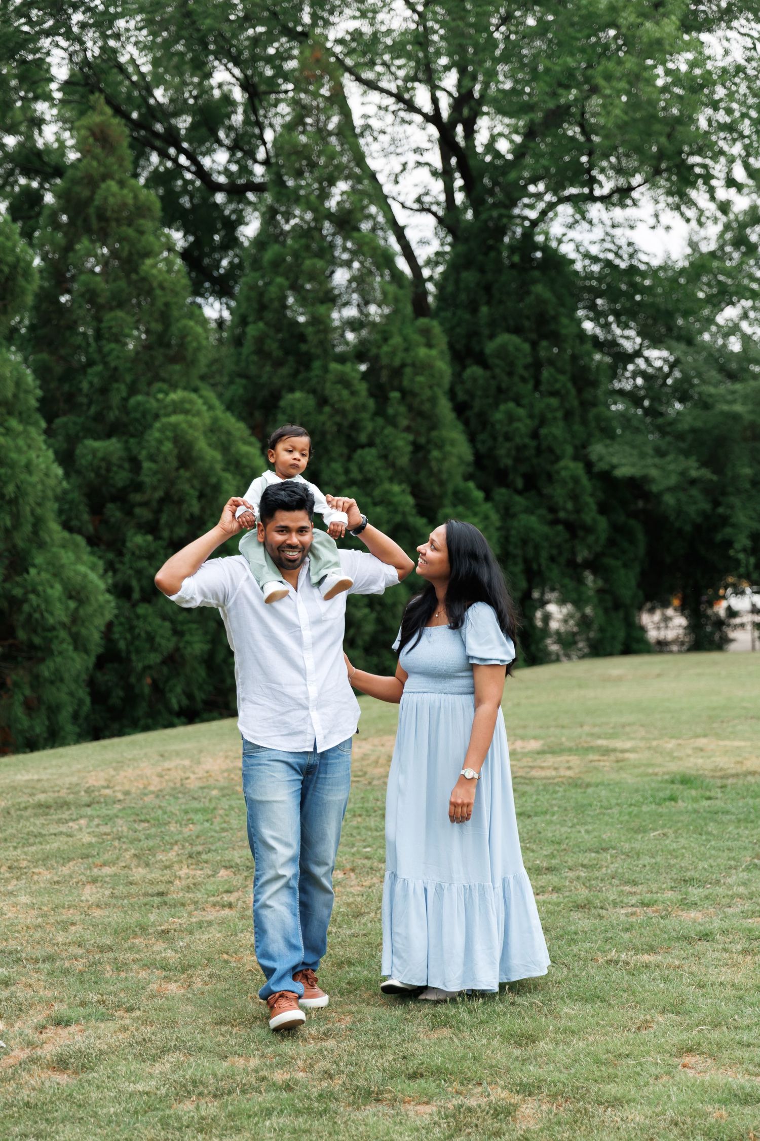 A smiling family of three poses on a sunny grassy lawn as a father carries his toddler on his shoulders beside the mother in a blue dress.