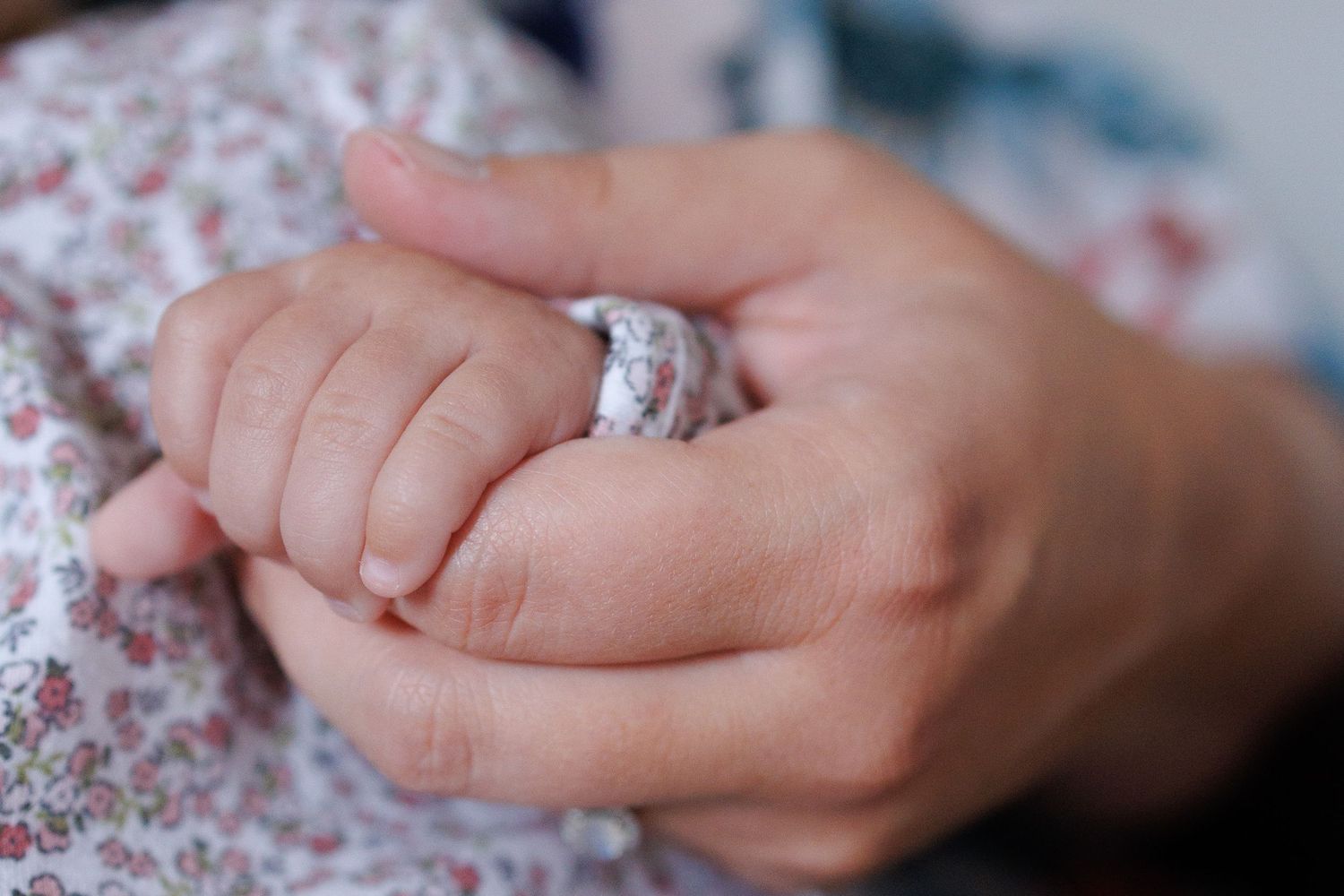 Close-up of an adult hand gently holding a sleeping newborn baby's tiny hand wrapped in a soft floral blanket.