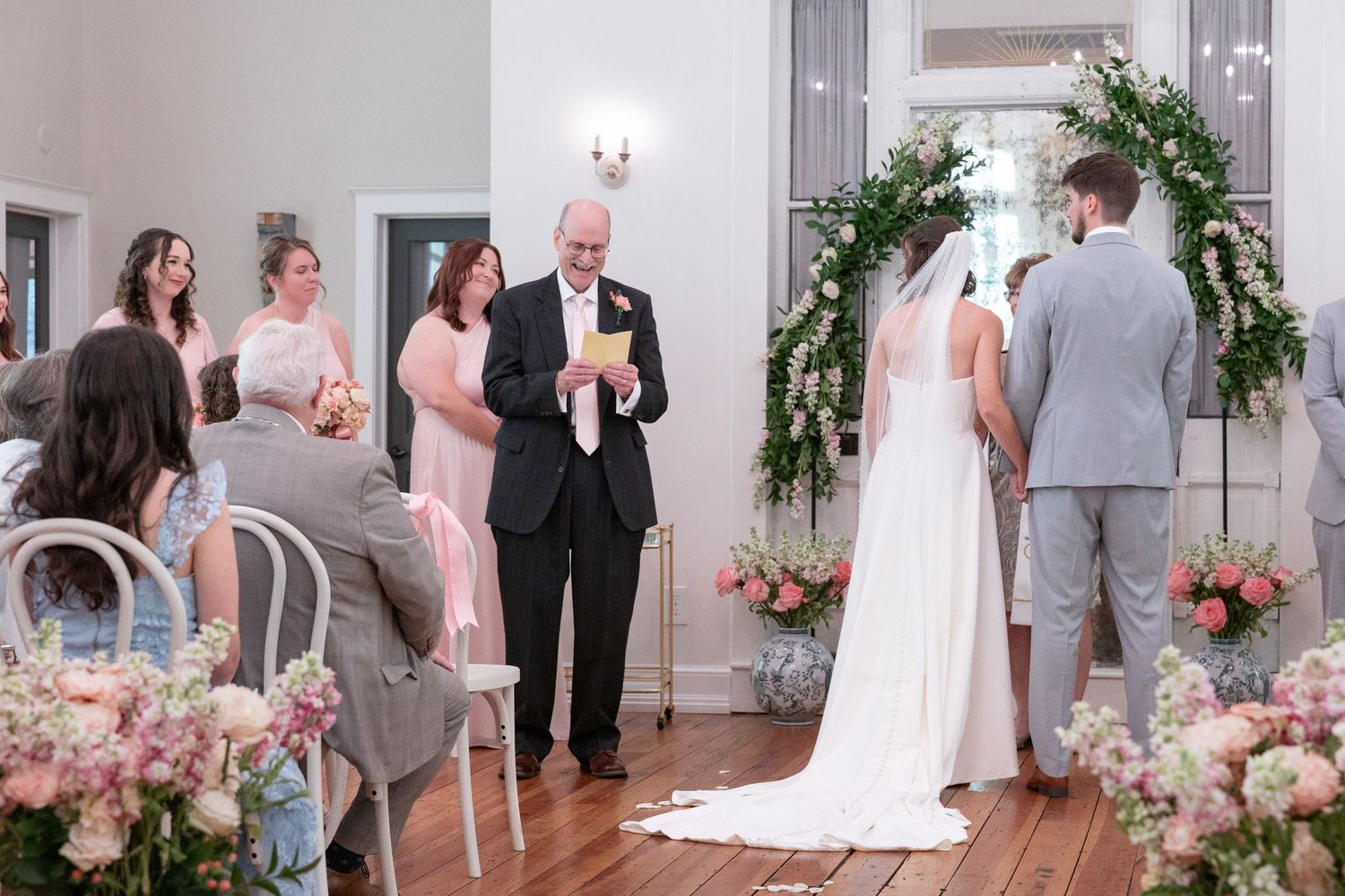 Bride and groom stand before an officiant during an indoor wedding ceremony with floral garland arch, pink bouquets, and seated guests.
