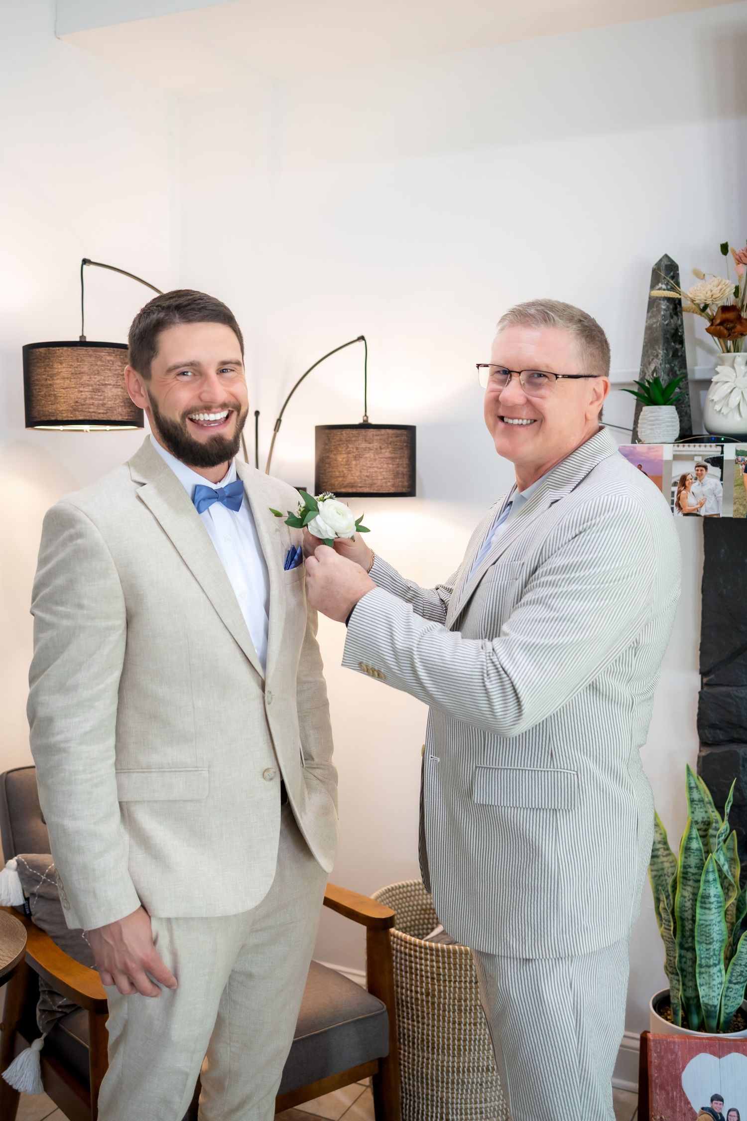 Smiling groom in a tan suit and blue bow tie has a white boutonniere pinned to his lapel by an older man in glasses.
