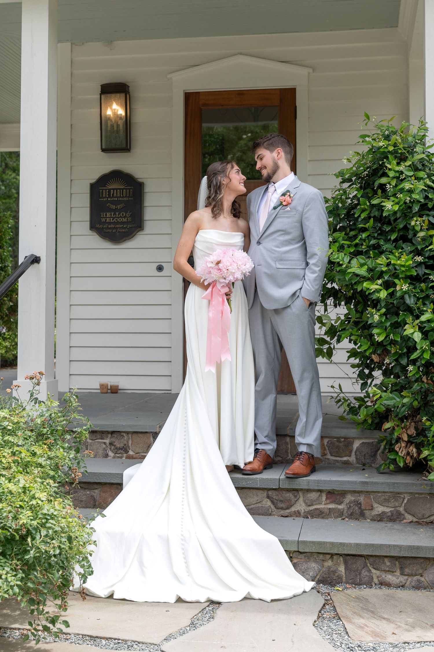 Bride in a white strapless gown with a long train holds a pink bouquet and smiles at her groom in a gray suit outside a white building.