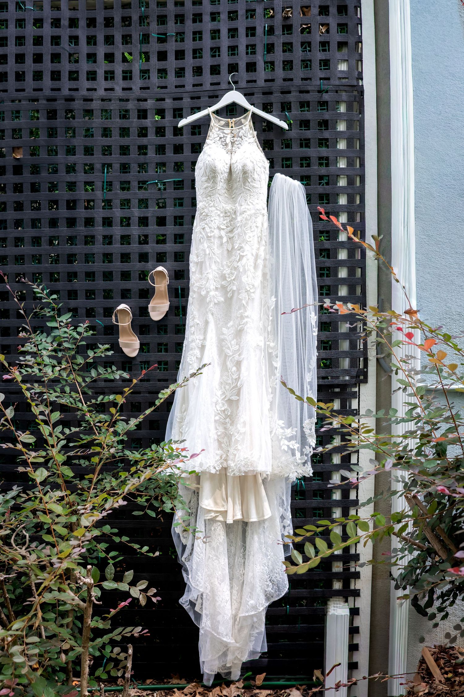 A lace wedding dress with a long veil hangs on a dark lattice wall beside a pair of nude heeled sandals.