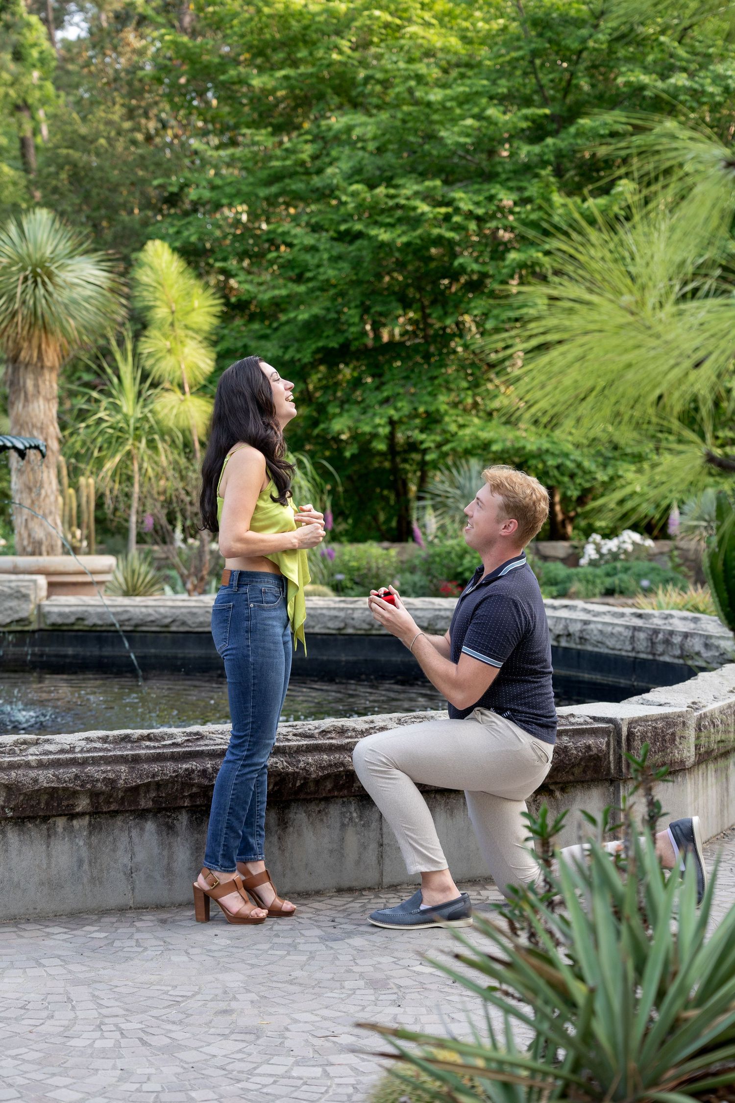Man kneeling on one knee proposing to woman with ring box near garden fountain surrounded by lush greenery.