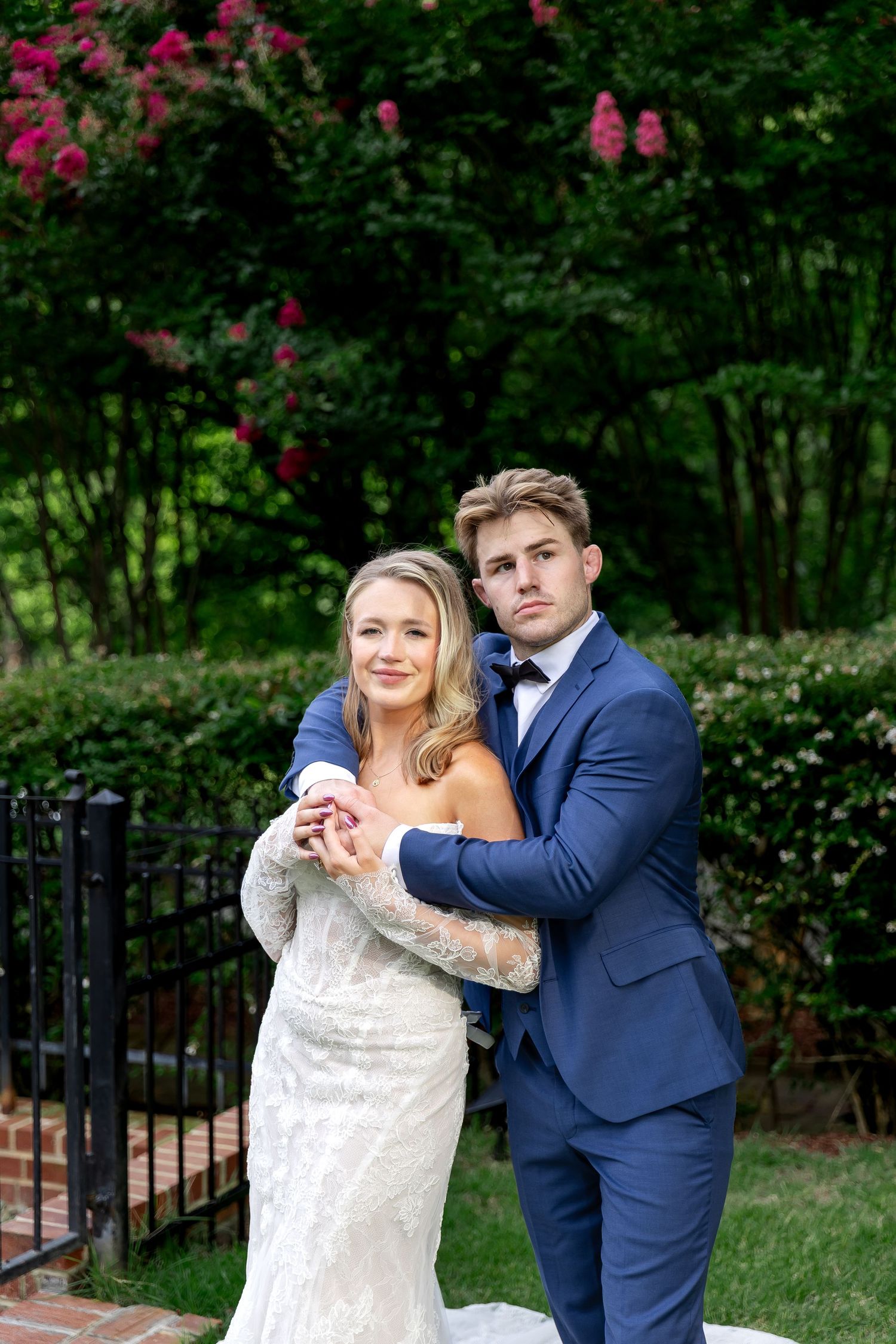 Bride and groom embrace outdoors in lush garden, wearing white gown and navy suit with bow tie.