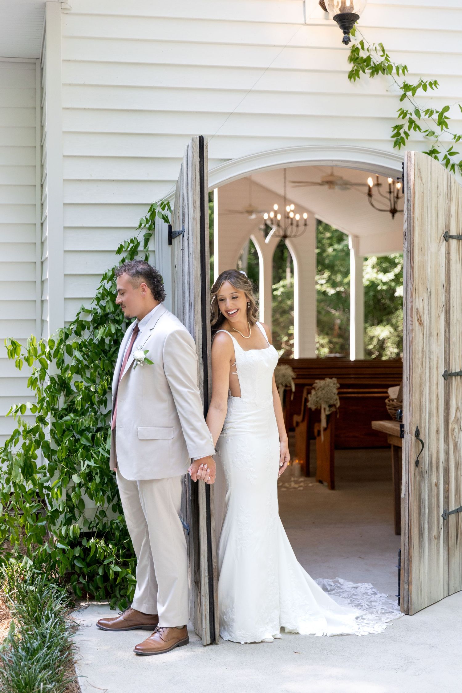 Bride and groom hold hands on opposite sides of an open wooden door in a first touch moment outside a white chapel with pews.
