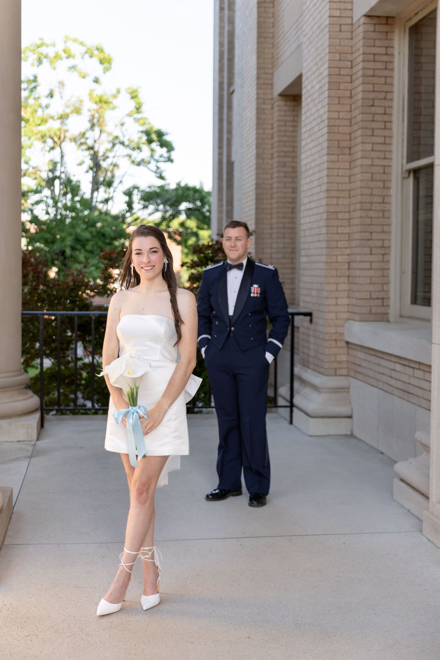 Smiling bride in short white dress holding calla lily bouquet with Air Force groom standing behind her outdoors.