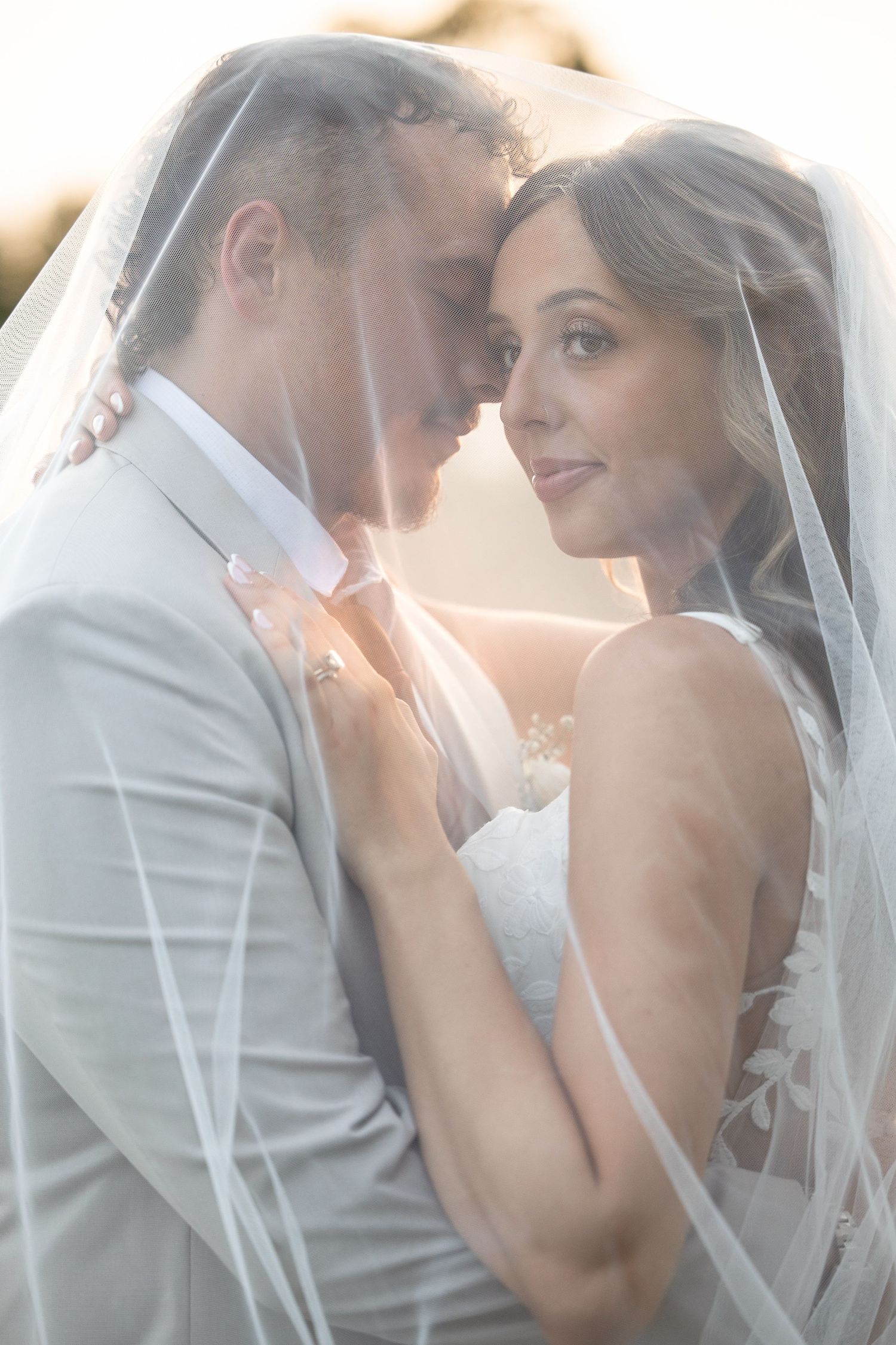 Romantic bride and groom portrait under a flowing veil with soft golden backlight at sunset.