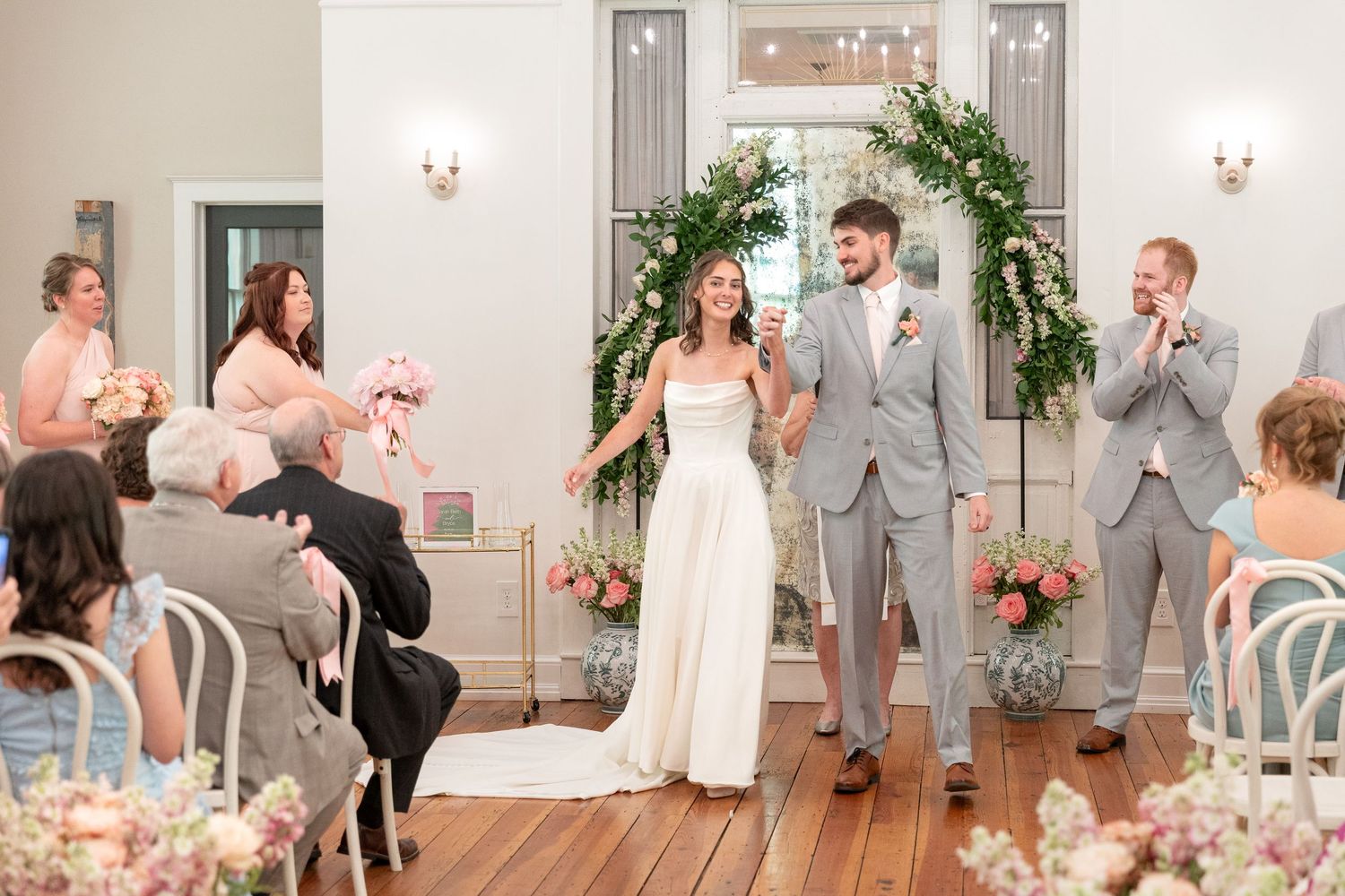 Smiling bride in a white gown and groom in a gray suit walk down the aisle past cheering wedding guests under a floral arch.
