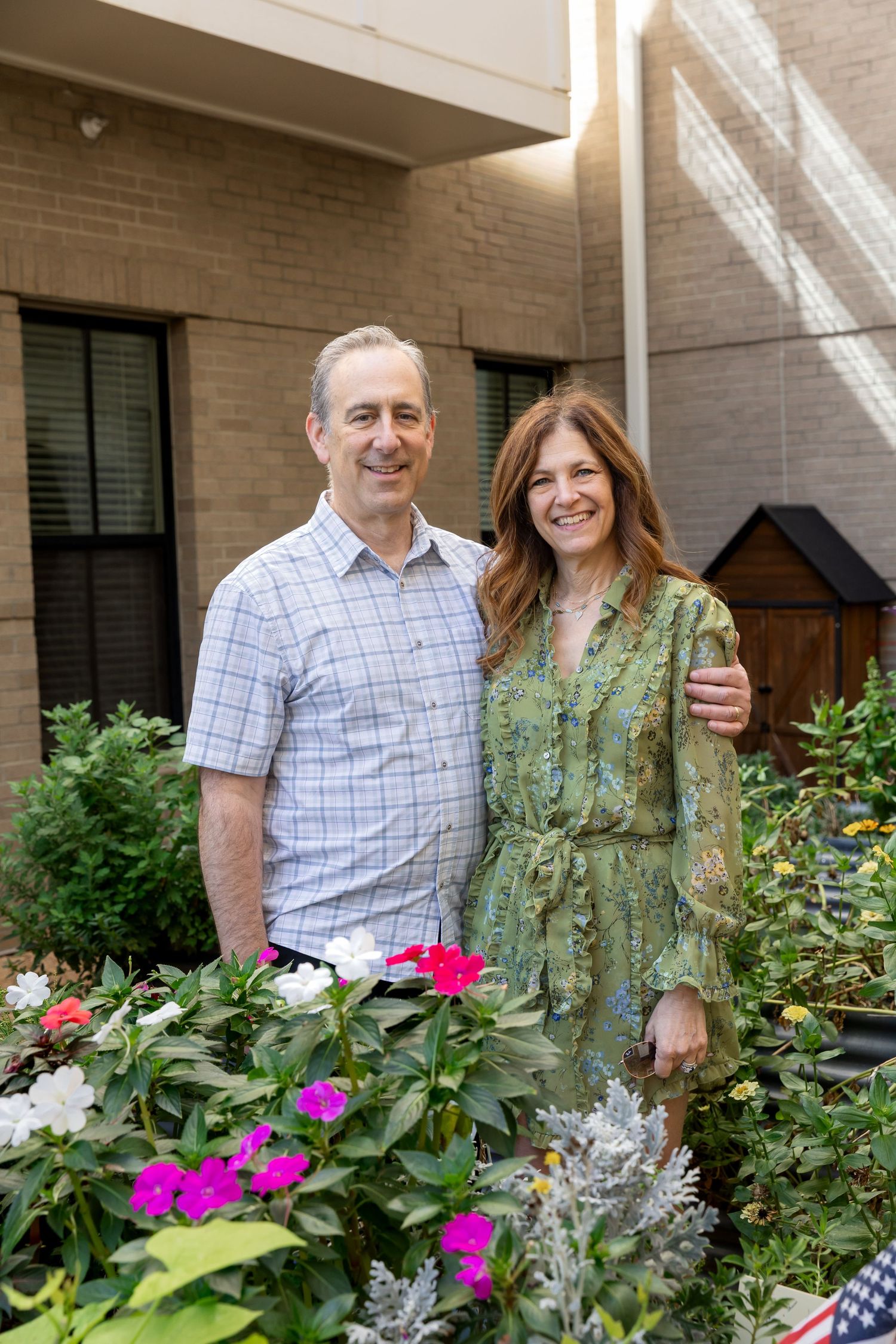 Smiling man in a plaid shirt and woman in a green floral dress pose together outdoors beside pink and white flowers near a brick building.