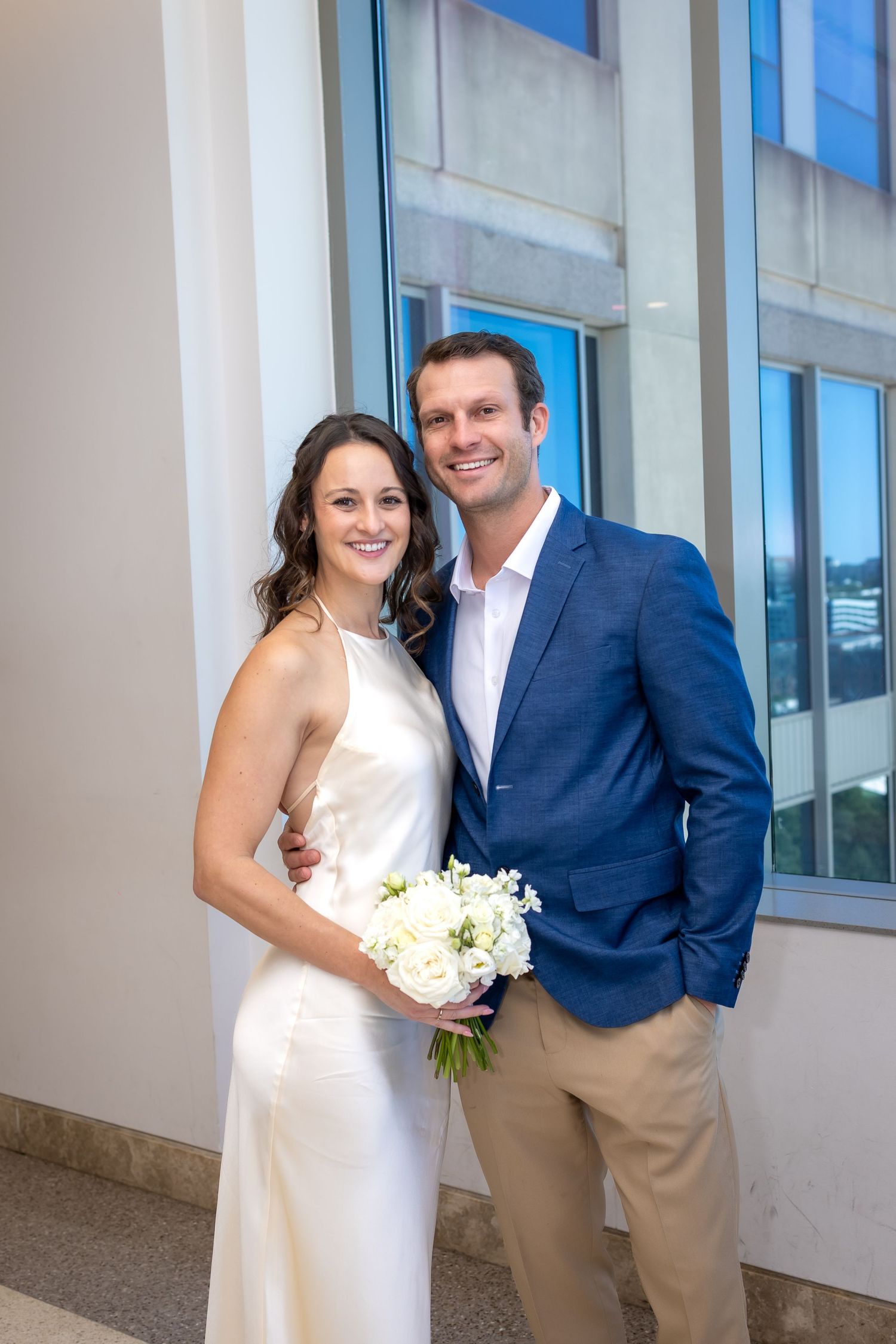 Smiling bride in white dress holding bouquet with groom in blue blazer posing together at wedding venue.