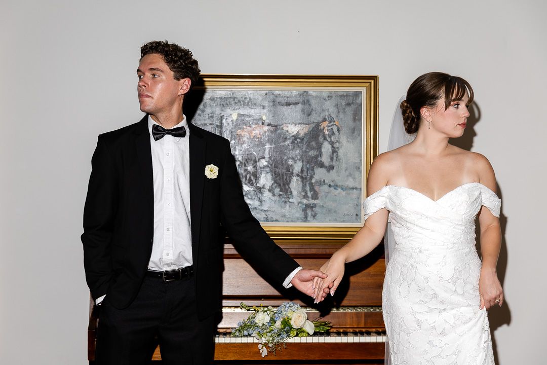 Bride and groom hold hands in front of a piano and framed painting, dressed in wedding attire.