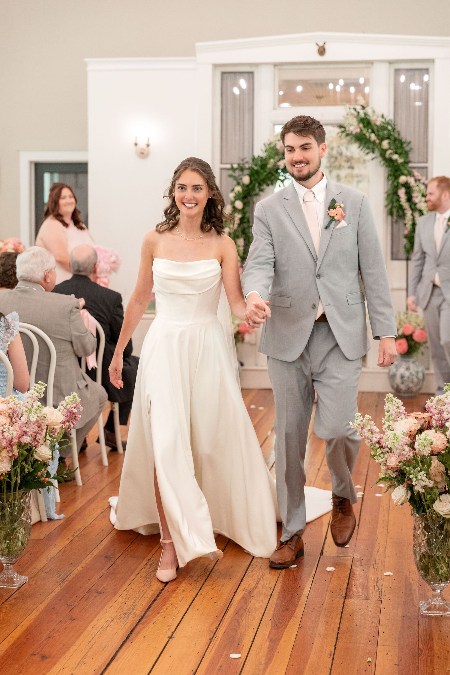 Smiling bride in a white satin gown and groom in a gray suit hold hands walking down the aisle past guests at their indoor wedding.