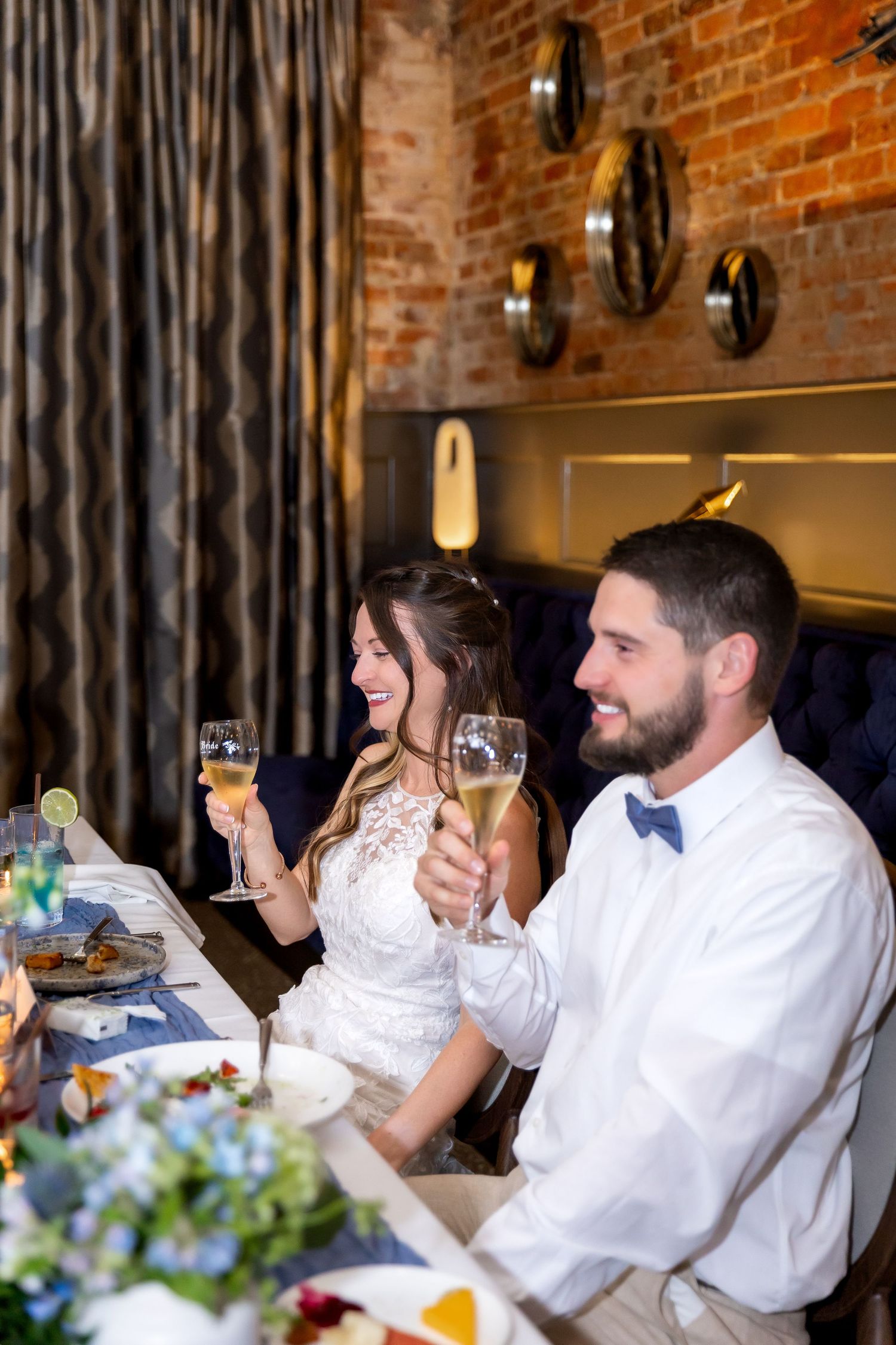 Smiling couple in wedding attire raise champagne glasses in a toast at a reception table with floral centerpieces and a brick wall.