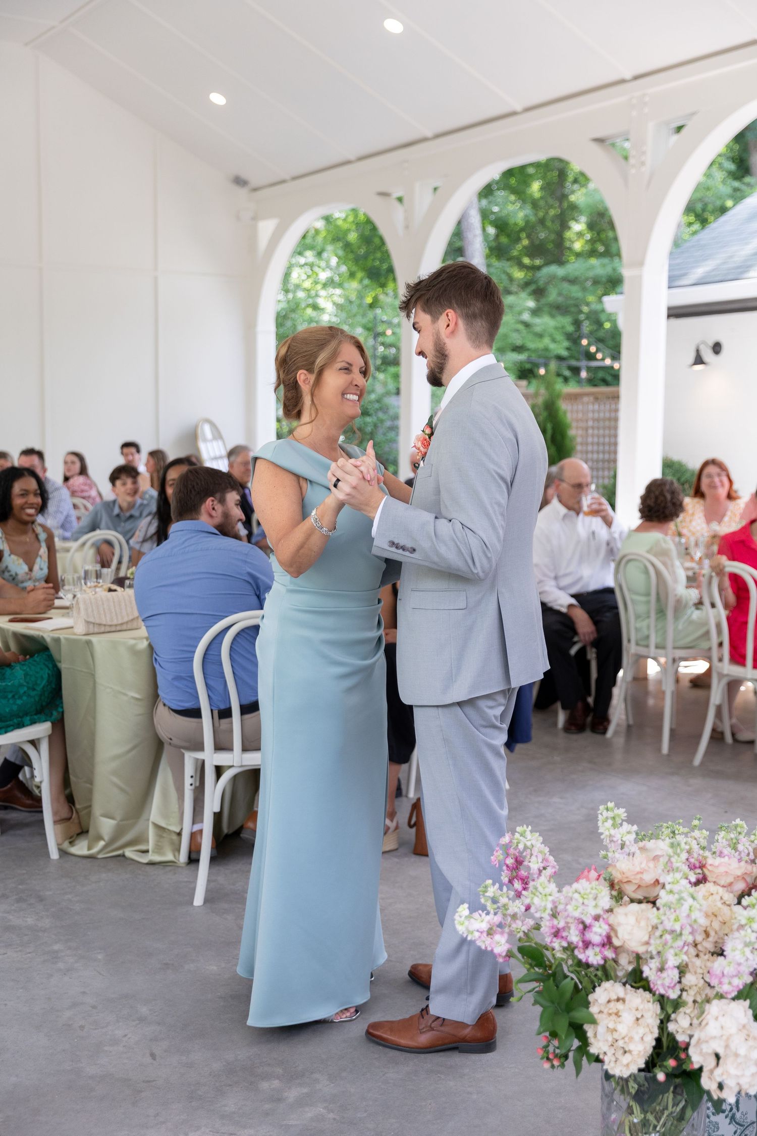 Groom in gray suit shares a mother-son dance under a white pavilion at an outdoor wedding reception.