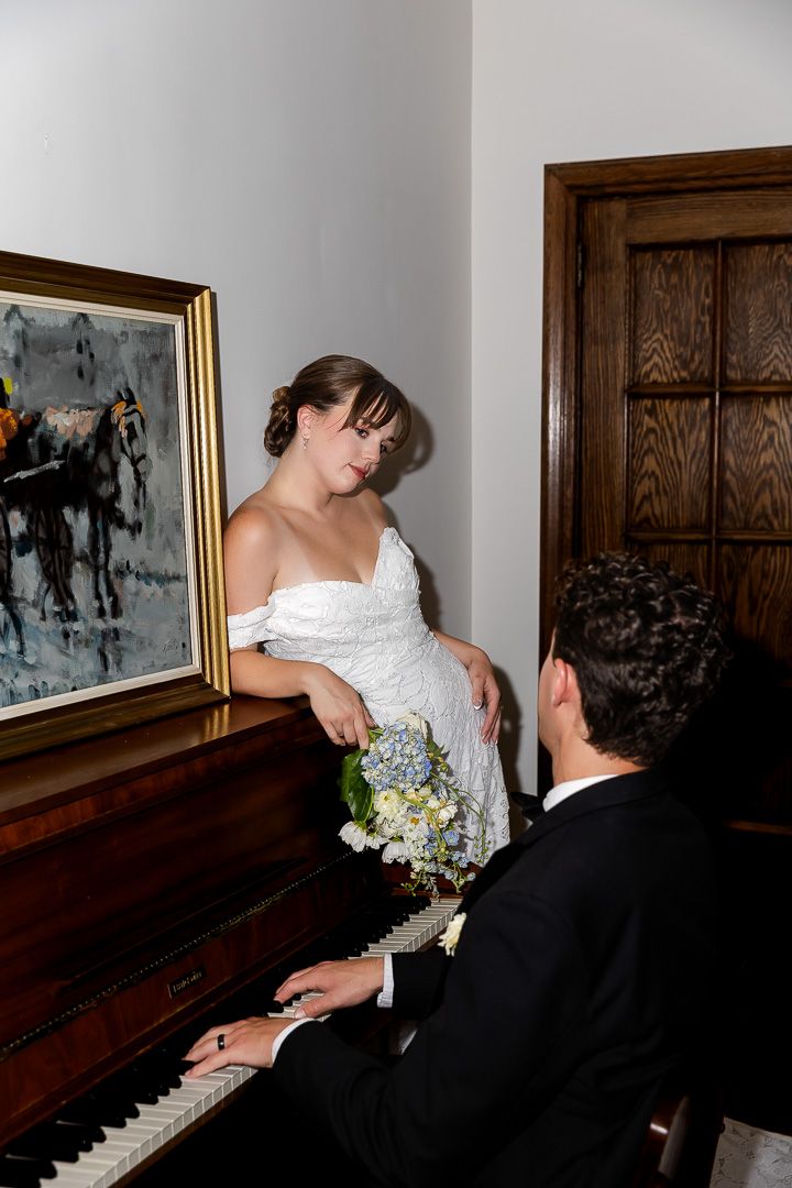 Bride in white off-shoulder gown leans on piano watching groom play in an elegant wood-paneled room.