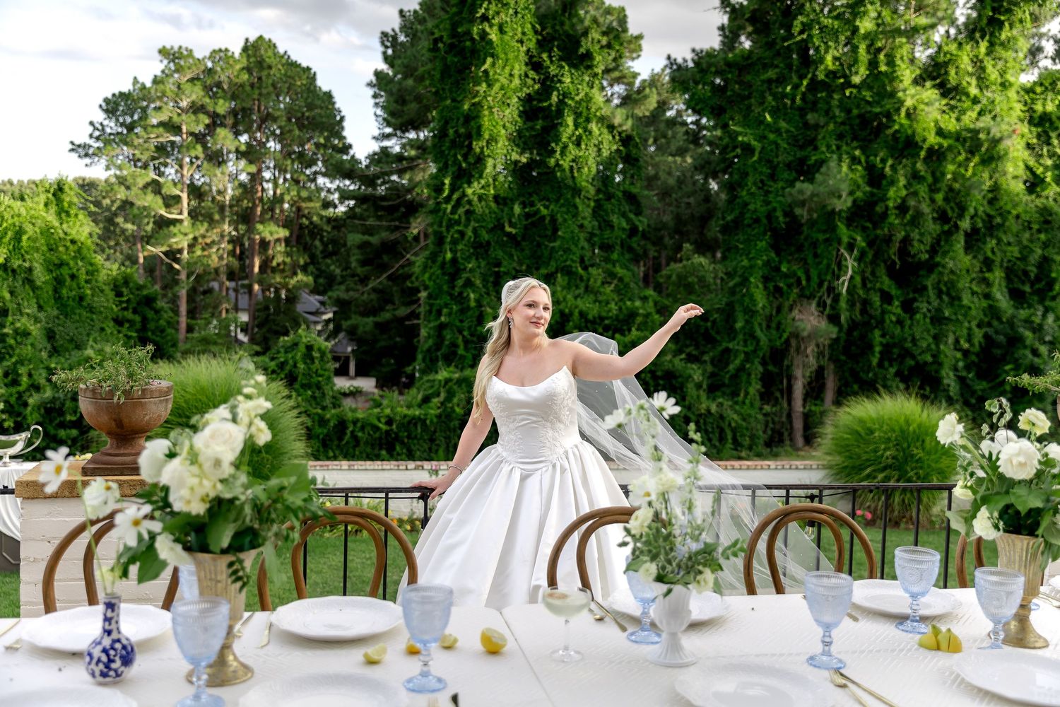 Bride in white gown gesturing at elegant outdoor wedding reception table with floral arrangements and lush greenery.