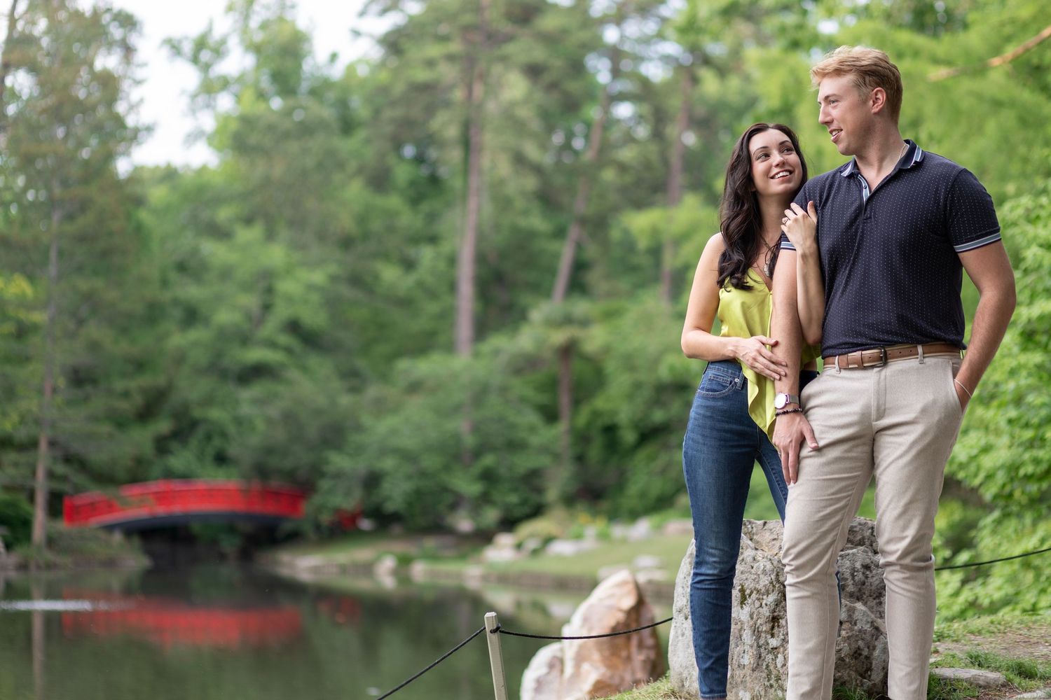 Couple standing by a serene pond with a red bridge and lush green trees in the background.