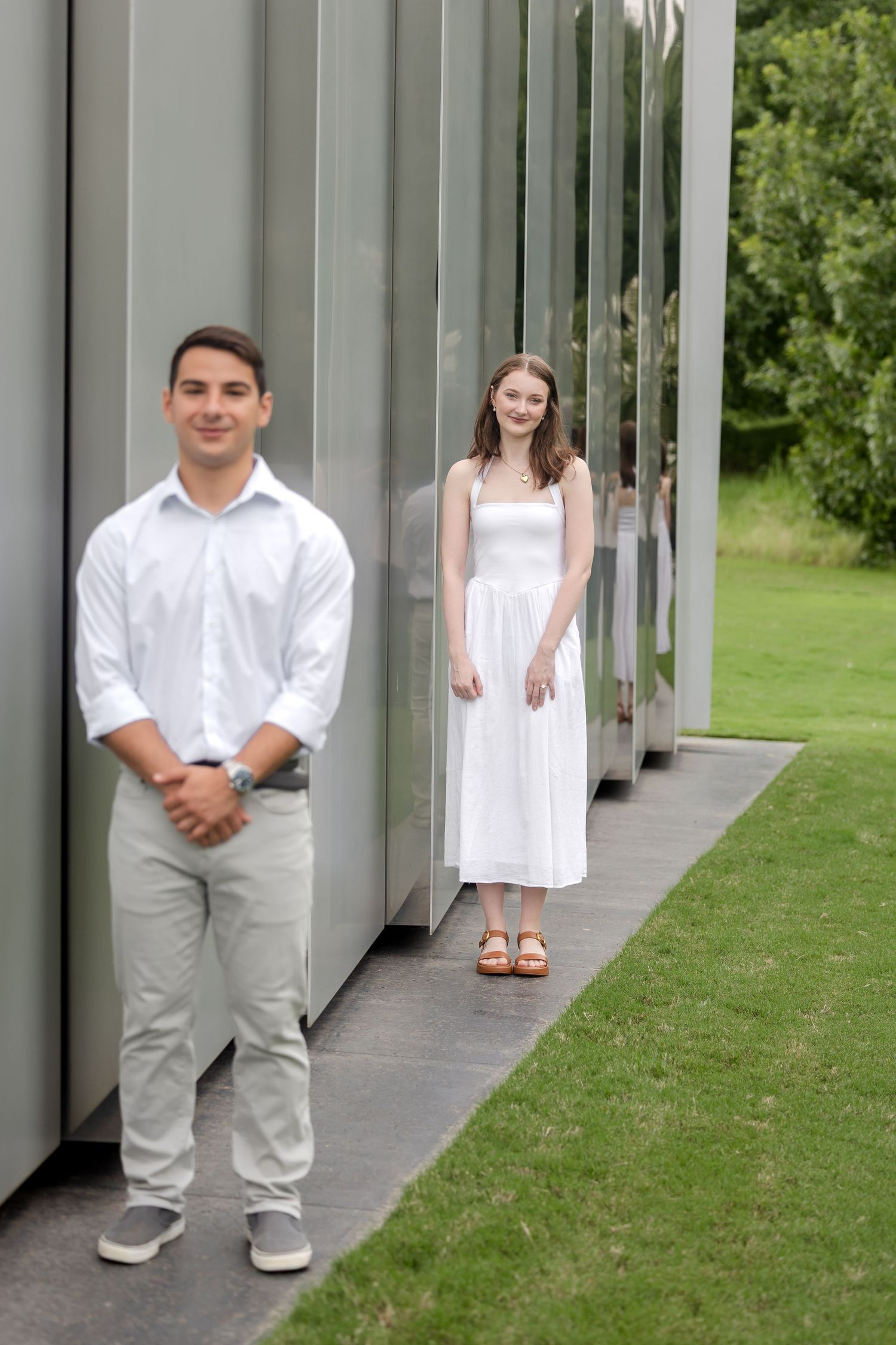 A young man and woman in white outfits pose near modern mirrored panels surrounded by green grass.