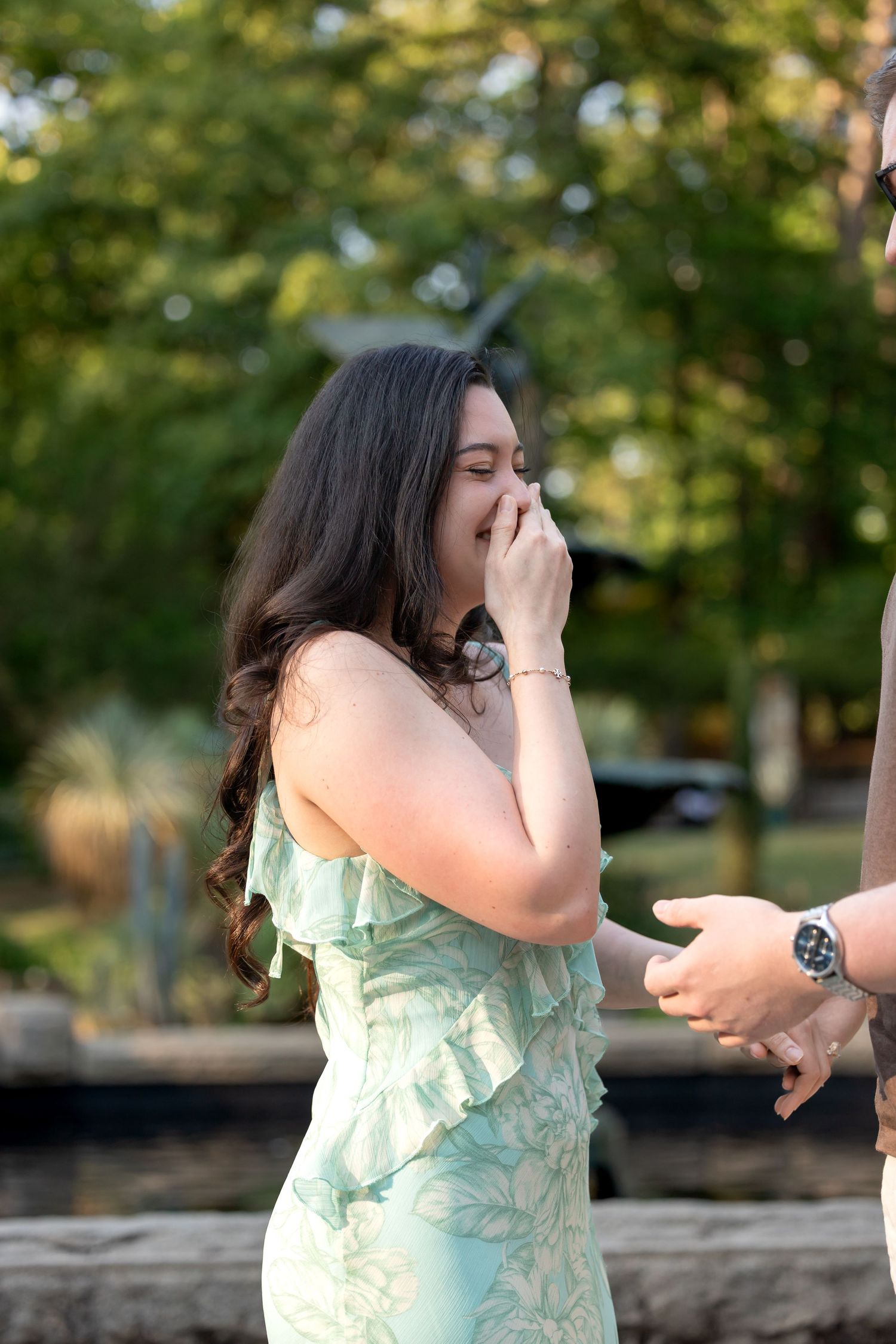 Woman in mint green dress covering her mouth in surprise during an outdoor marriage proposal in a garden setting.