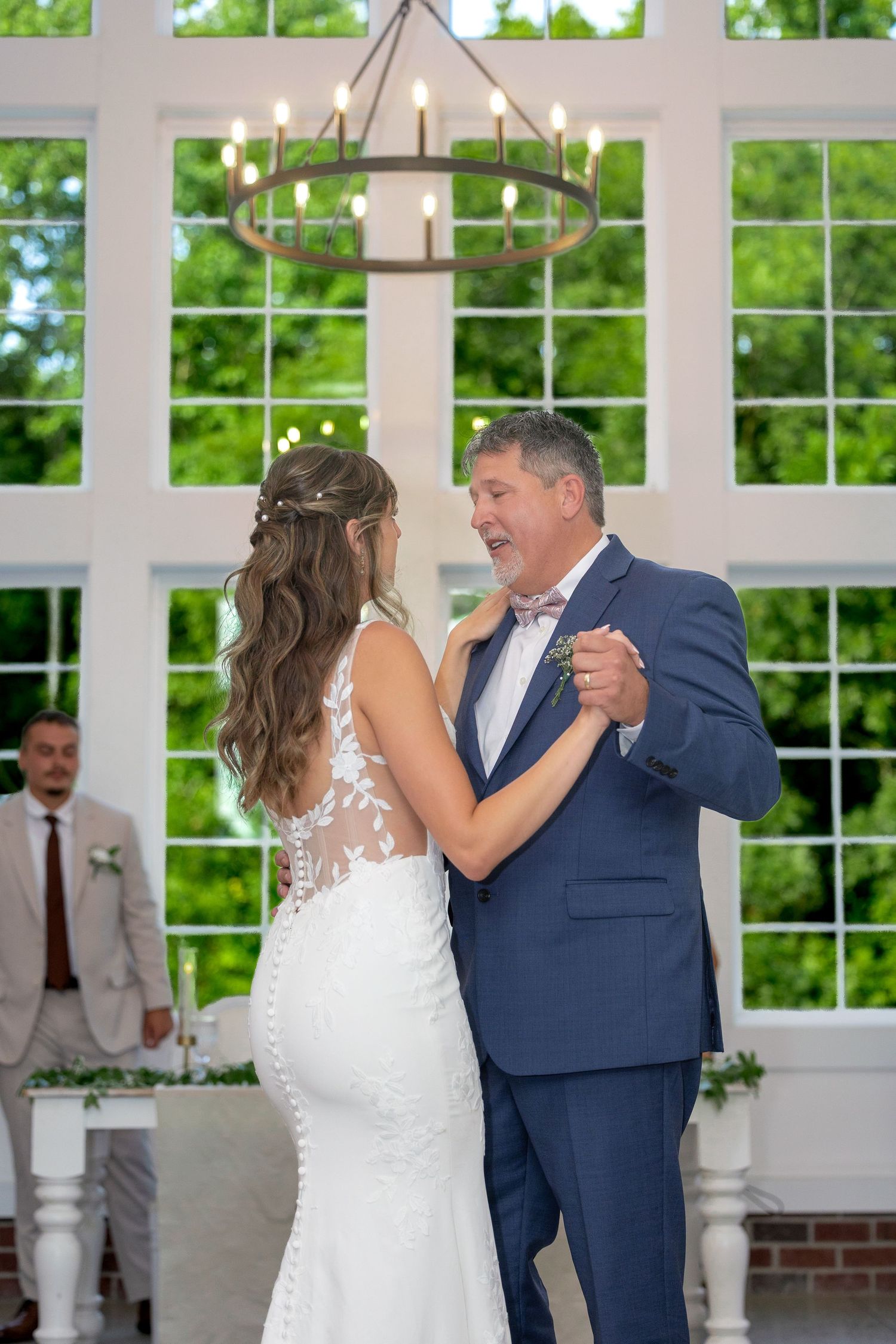 Bride in a lace wedding gown dances with a man in a blue suit and bow tie beneath a candle chandelier in a bright, window-lined venue.
