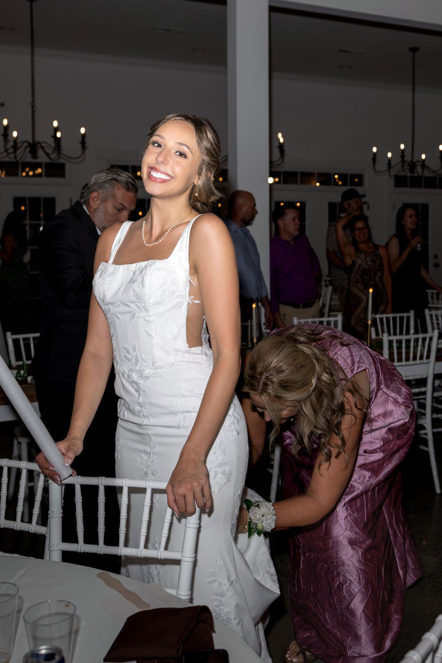 Smiling bride in a fitted white lace gown stands beside white chairs at an indoor wedding reception while guests mingle under chandeliers.