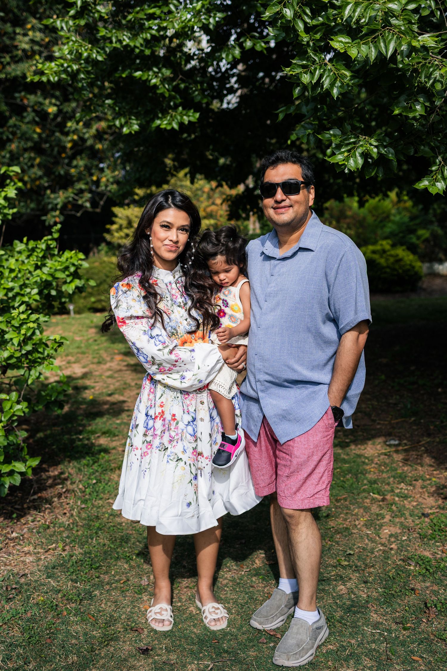 Young couple standing outdoors under a tree holding their toddler daughter during a family portrait session.