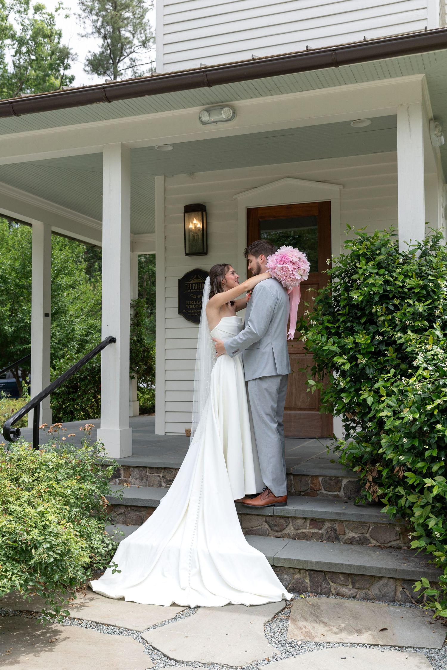 Bride in a long white gown and veil embraces groom in a gray suit holding a pink bouquet on the stone steps of a white porch.