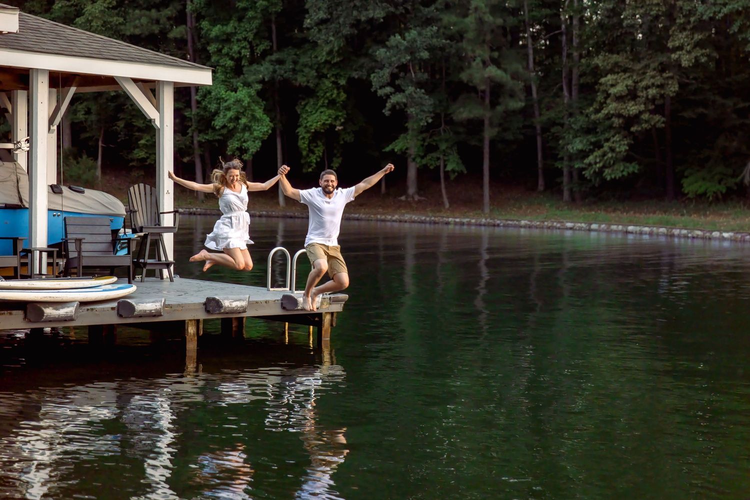 Smiling couple holding hands and jumping off a wooden lakeside dock into calm green water, surrounded by lush forest trees.