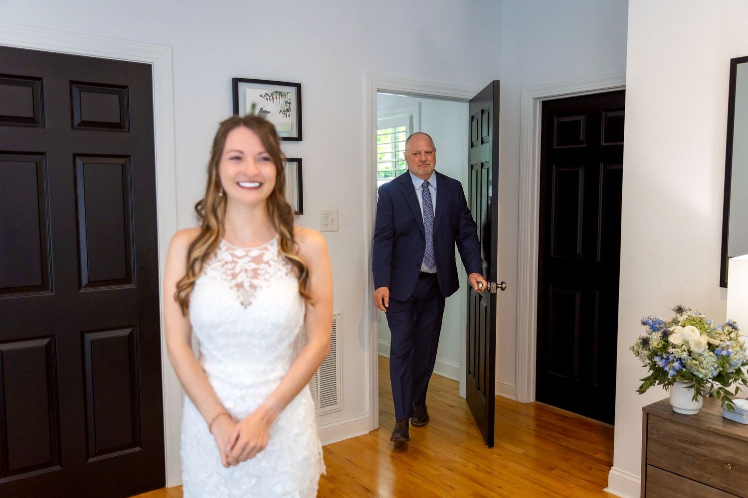 Bride in white lace dress smiles as groom in navy suit approaches through doorway during wedding first look.