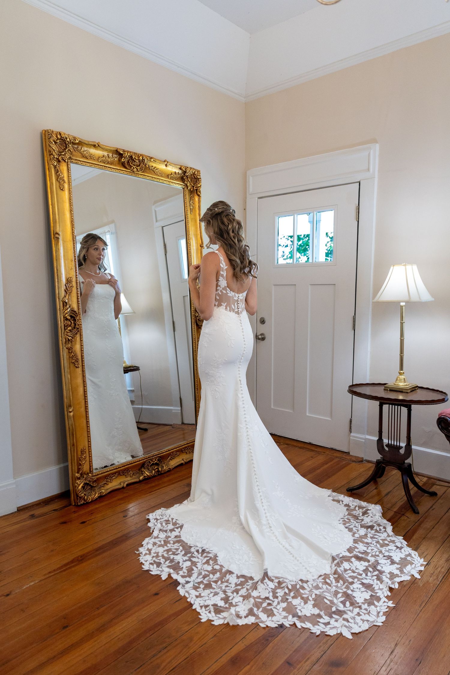 Bride in elegant lace mermaid wedding gown views her reflection in an ornate gold-framed floor mirror.
