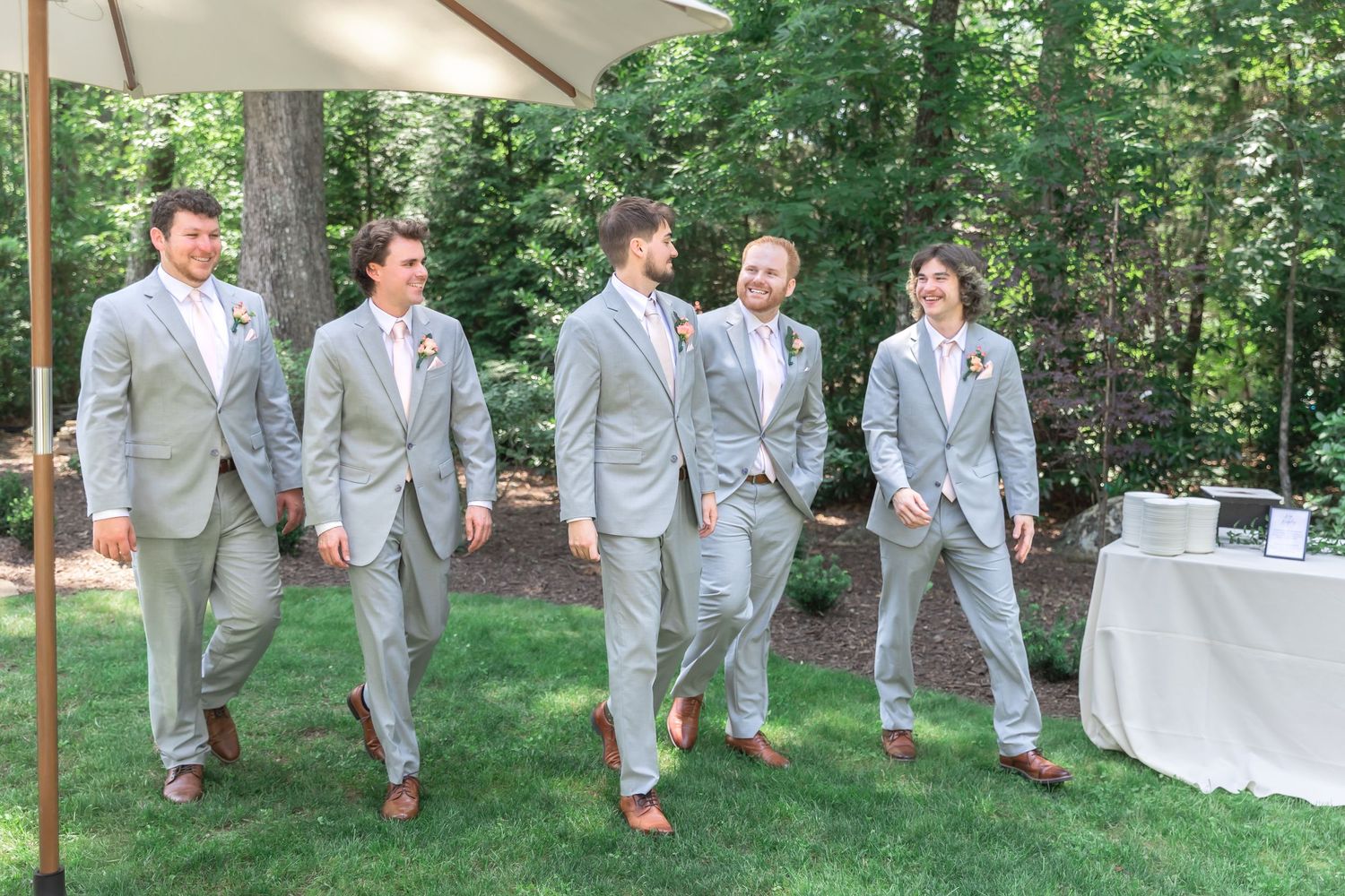 Five groomsmen in matching light gray suits walk together outdoors on green grass at a wedding celebration.
