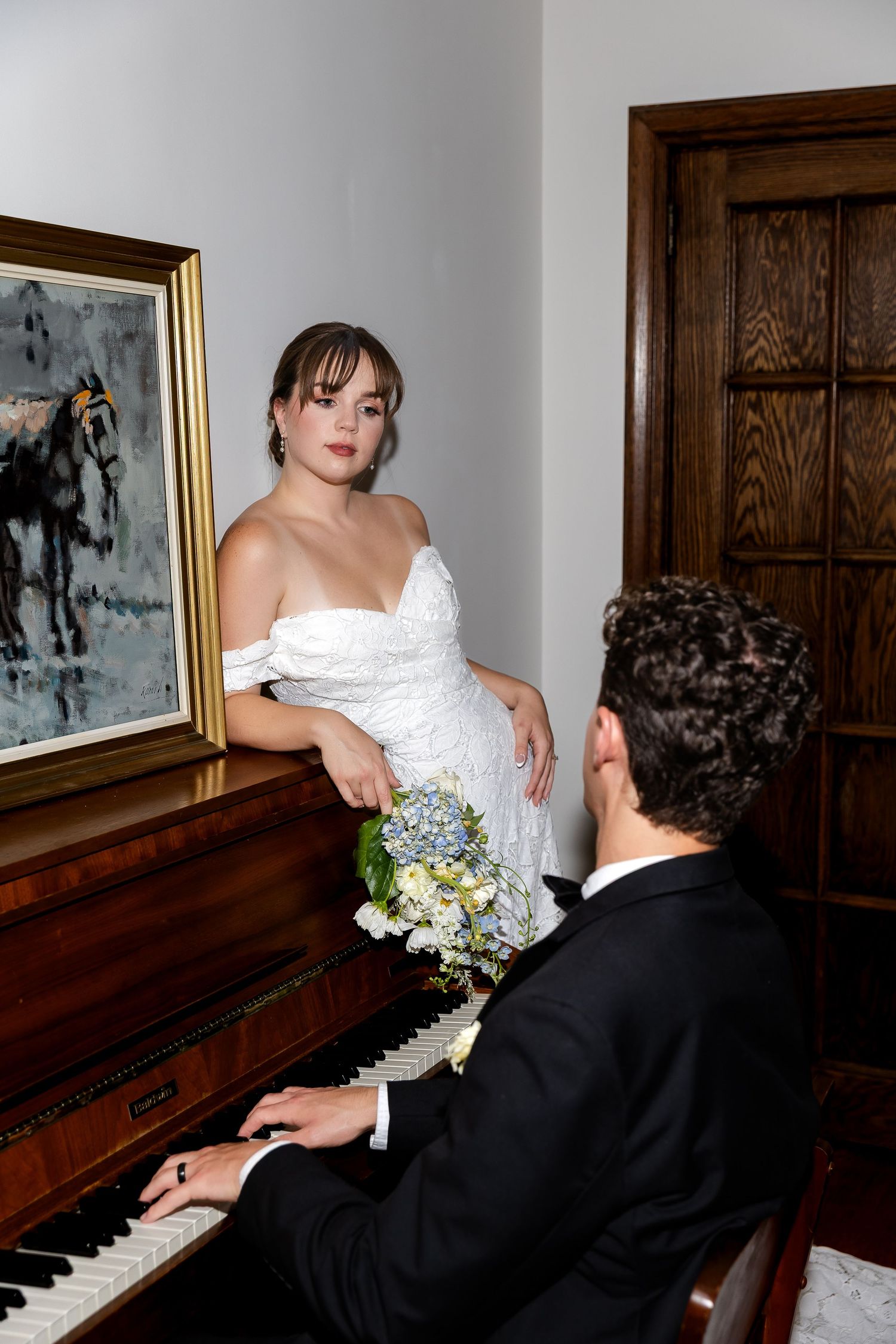 Bride in lace off-shoulder gown leans on piano while groom in tuxedo plays during wedding portraits.