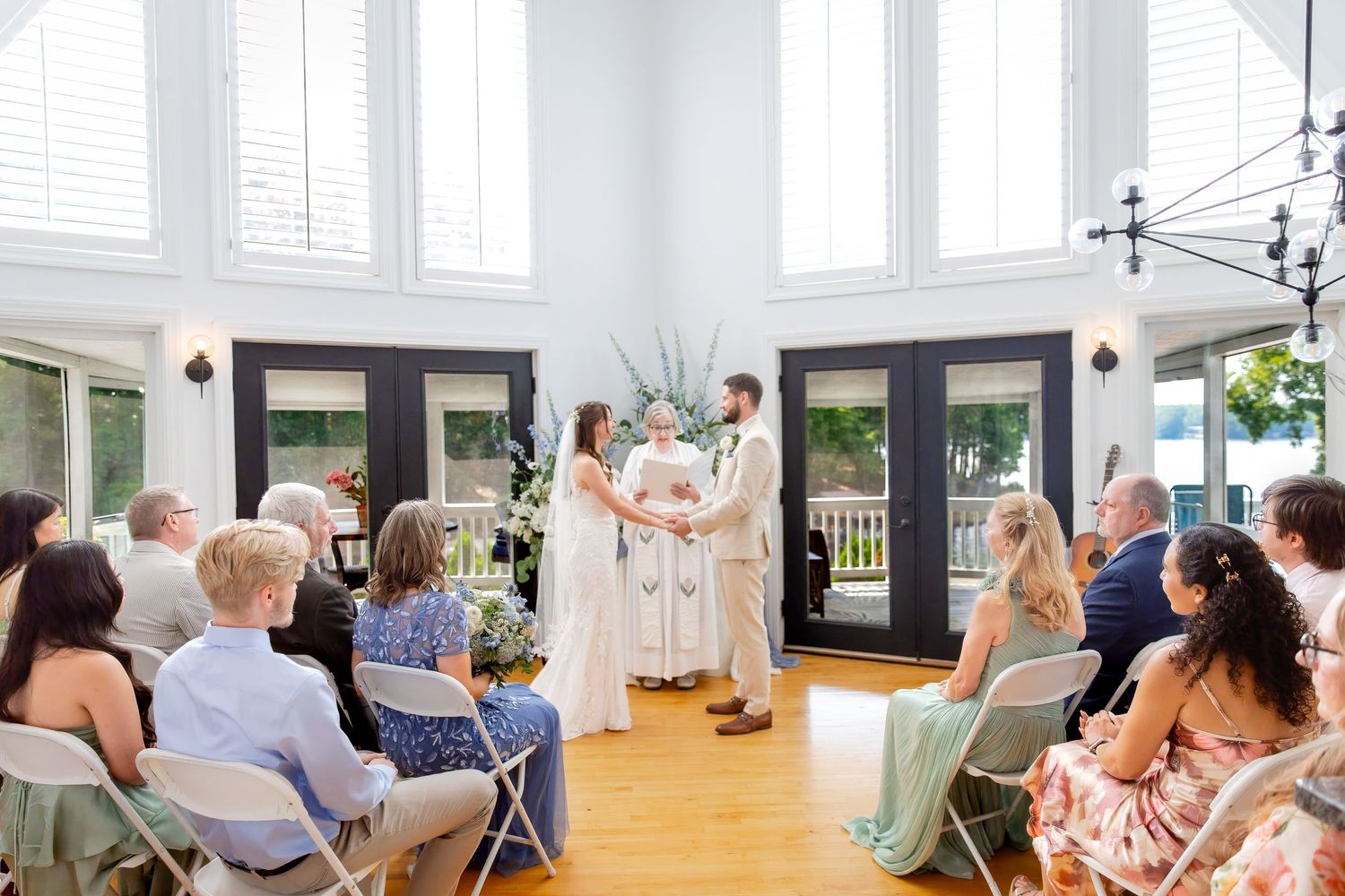Bride and groom exchange vows before an officiant during an intimate indoor wedding ceremony with seated guests and bright windows.
