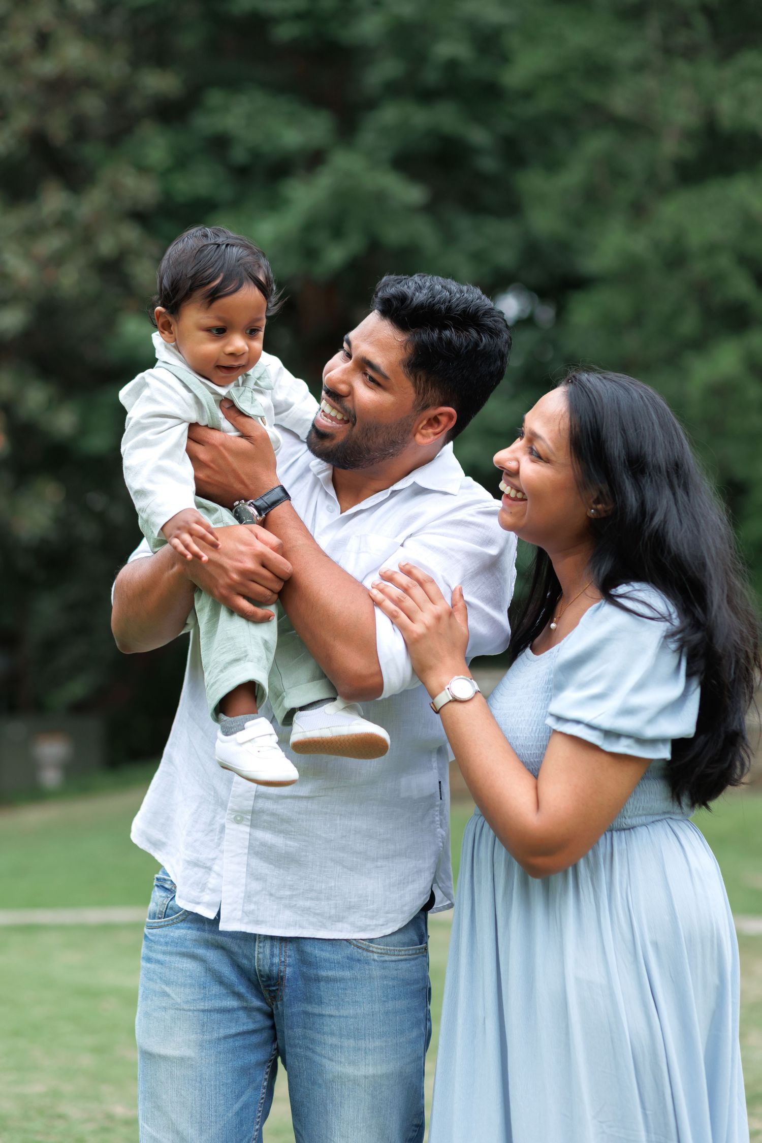 Smiling father holding a happy baby while a mother in a light blue dress hugs them close in a sunny green park.