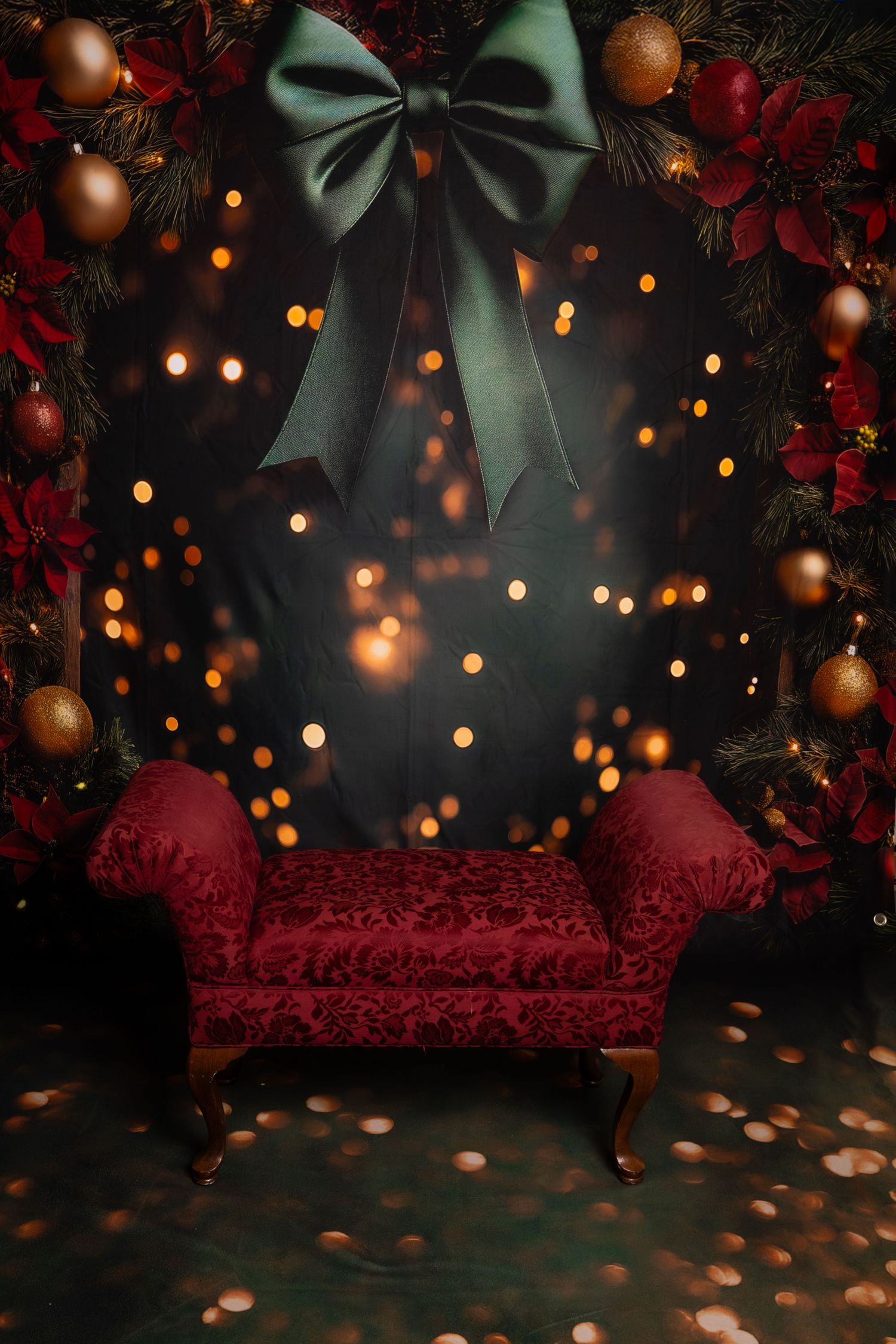 Red velvet chaise lounge against dark backdrop with Christmas lights and ornaments creating a festive holiday photo setting.