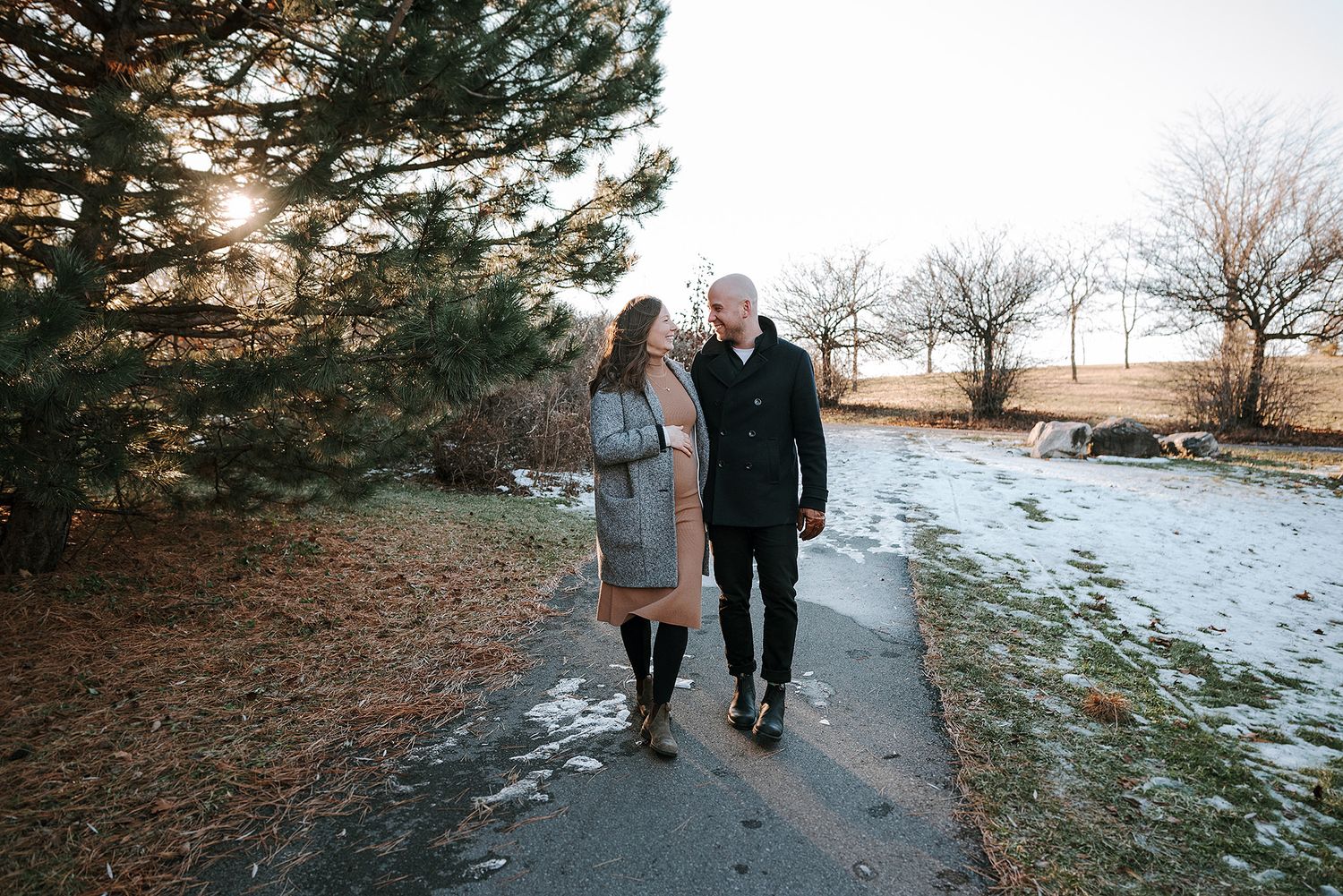 A couple walks together on a snowy winter path surrounded by bare trees and patches of snow on the ground.
