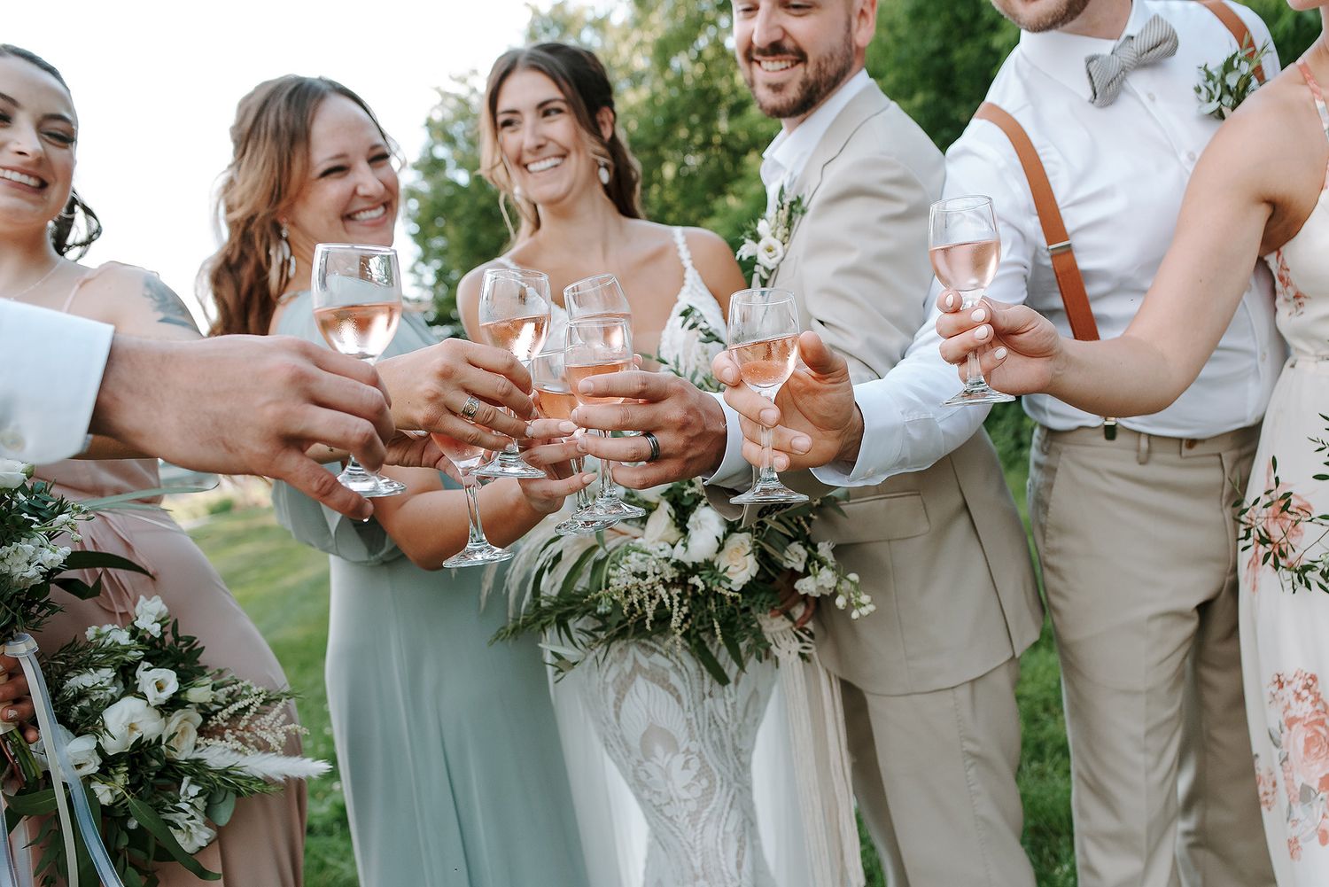 Wedding party in sage green dresses and tan suits toast with champagne glasses in an outdoor garden setting.