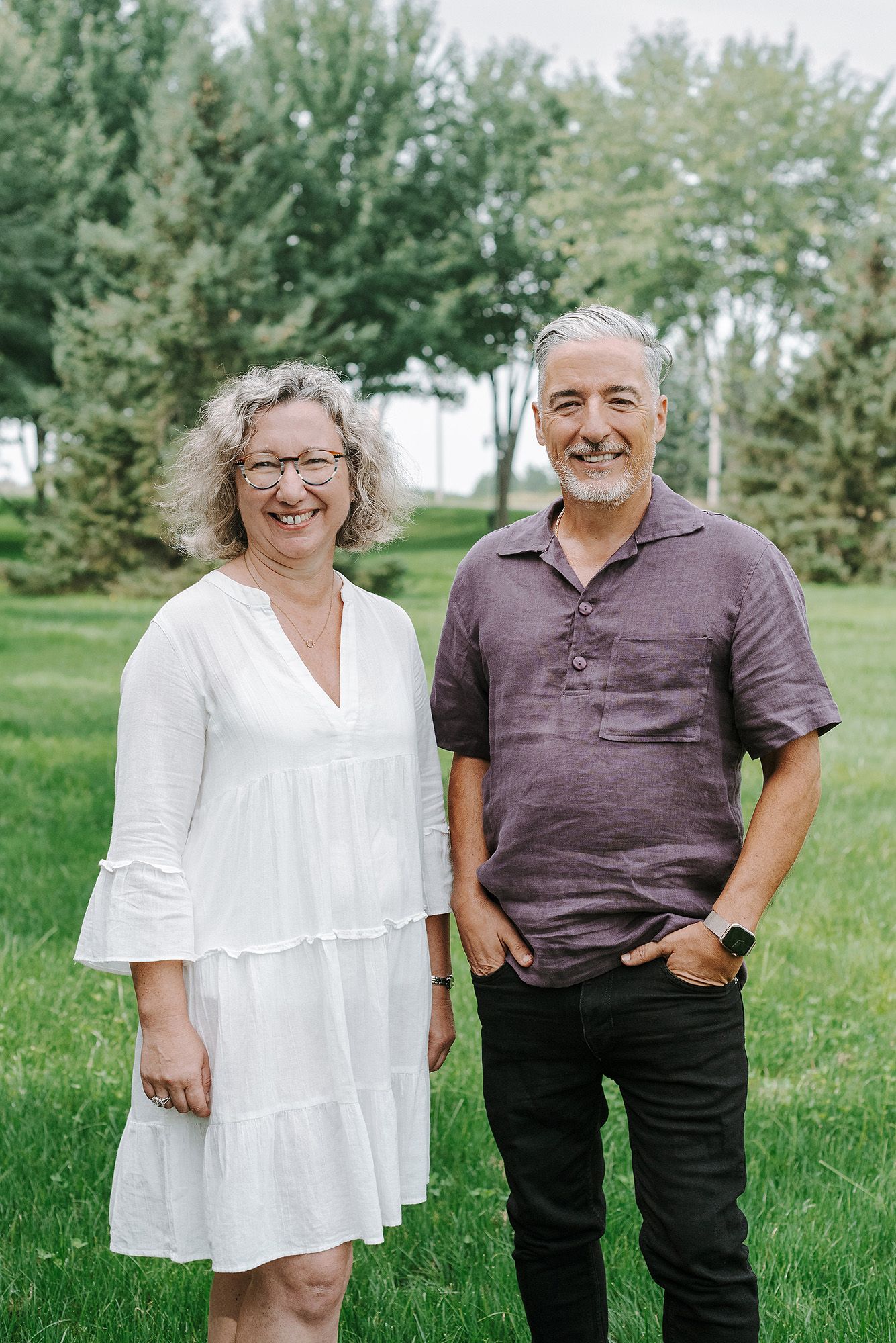 A couple stands together outdoors in a grassy field with trees in the background on a sunny day.