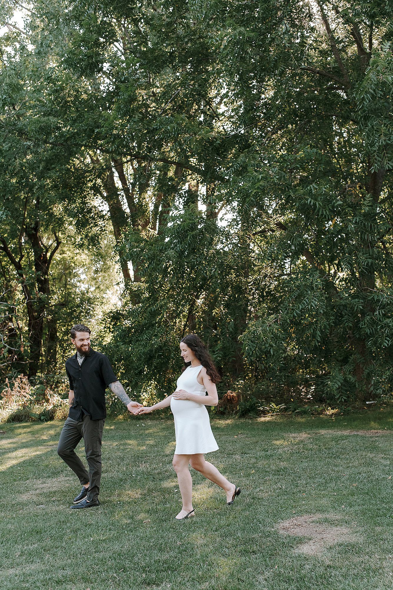 A couple playfully chases each other across a grassy hill with trees in the background.