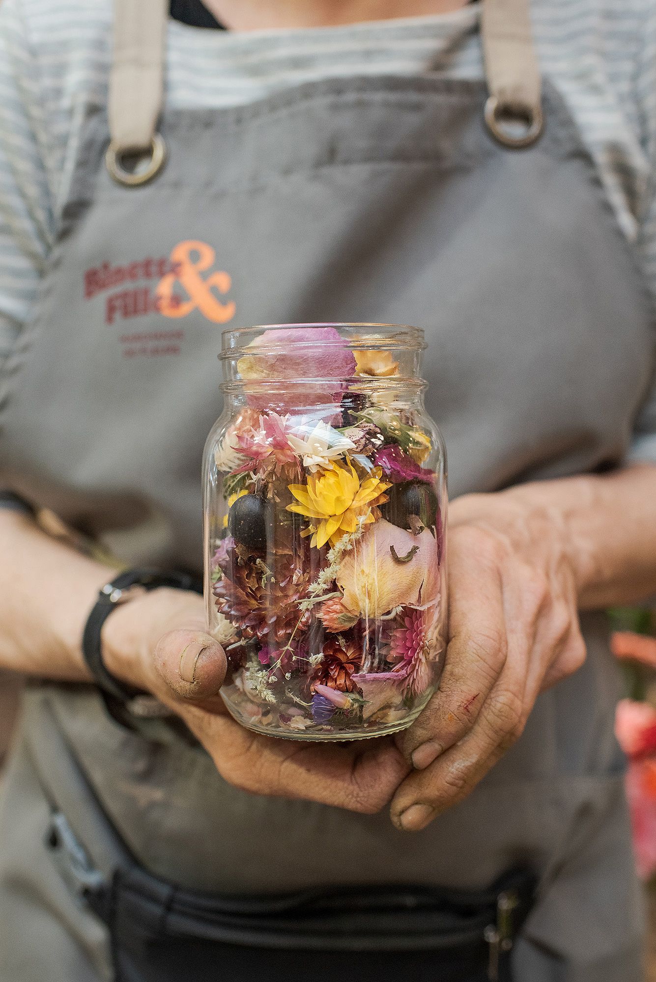 Glass jar filled with colorful dried flowers and petals being held against a gray apron background.